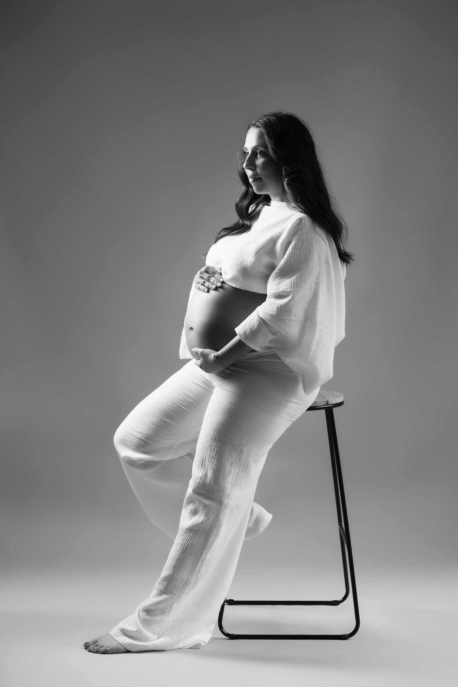Black and white maternity photo of pregnant woman in white outfit seated on stool on gray background.