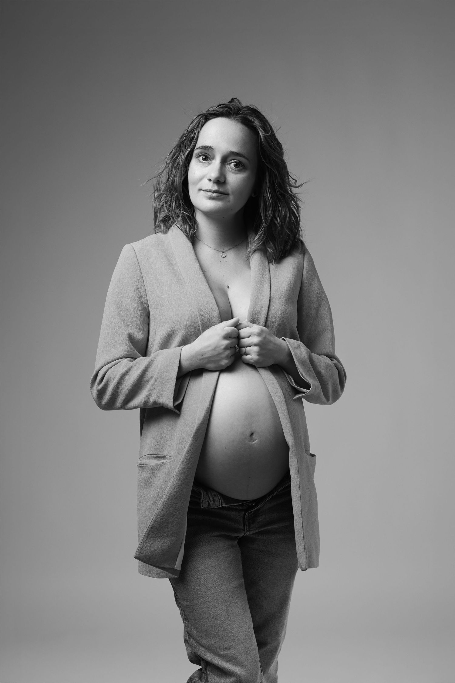 Black and white portrait of pregnant woman in blazer and jeans holding jacket open.