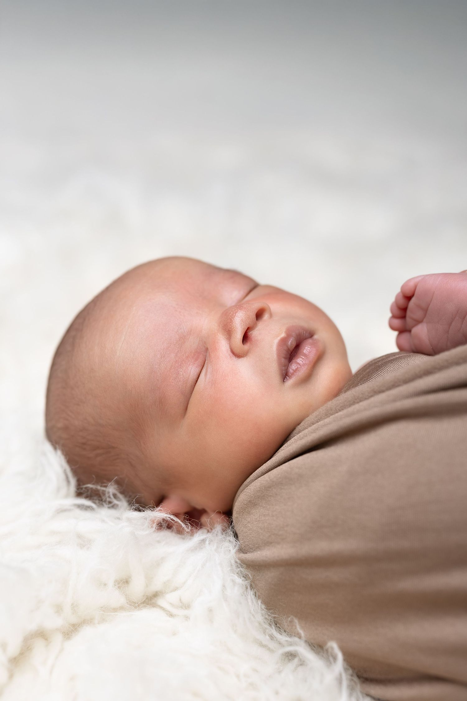 Peaceful newborn baby swaddled in a brown wrap sleeping on a soft white fluffy blanket. séance naissance chavanoz lyon