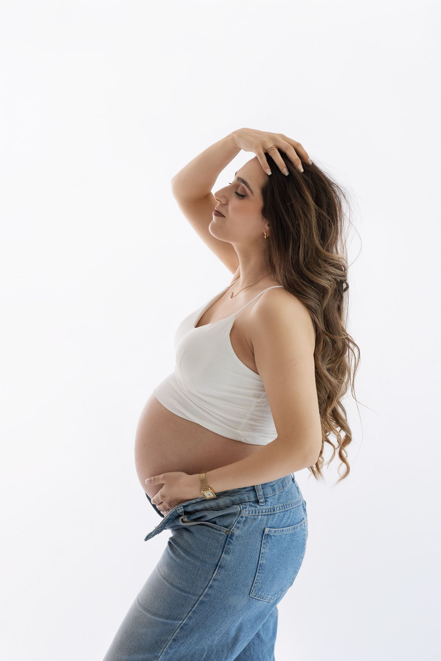 Pregnant woman in white crop top and jeans cradling her baby bump against a white background. photographe grossesse chavanoz lyon