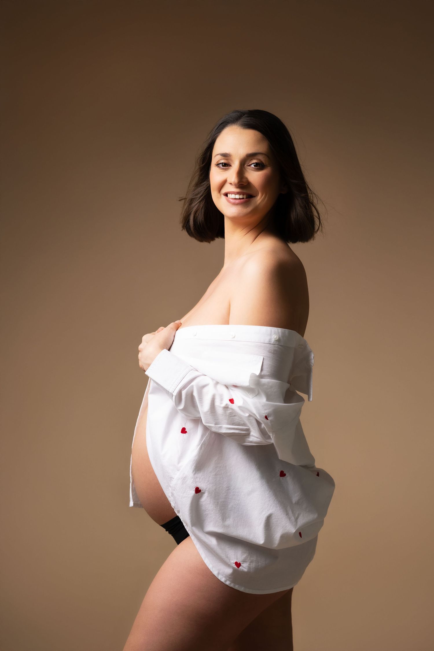 Smiling pregnant woman in white shirt with red hearts, posing with bare shoulders on beige background.