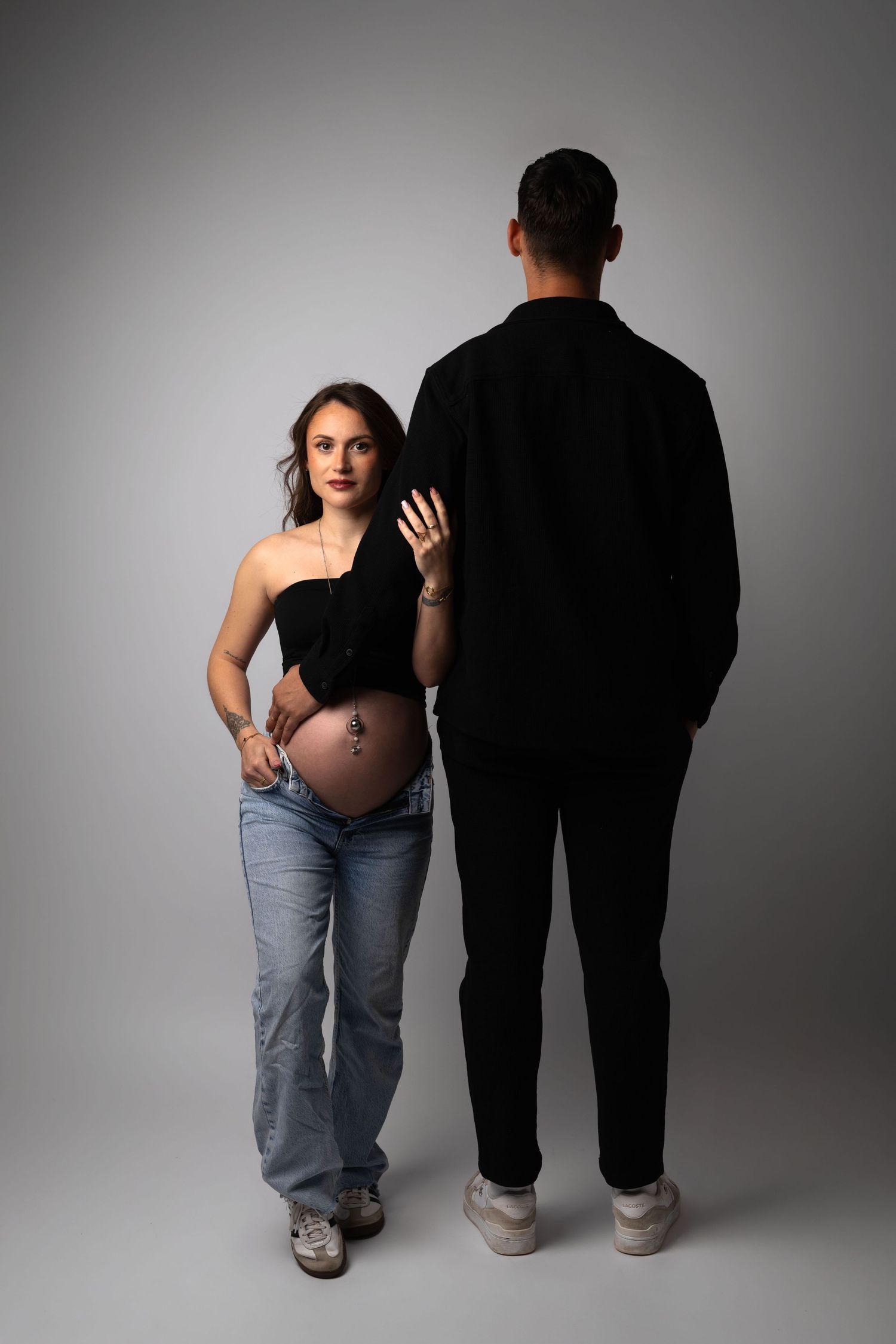Pregnant woman in black top and jeans poses with partner in black outfit against gray studio background.