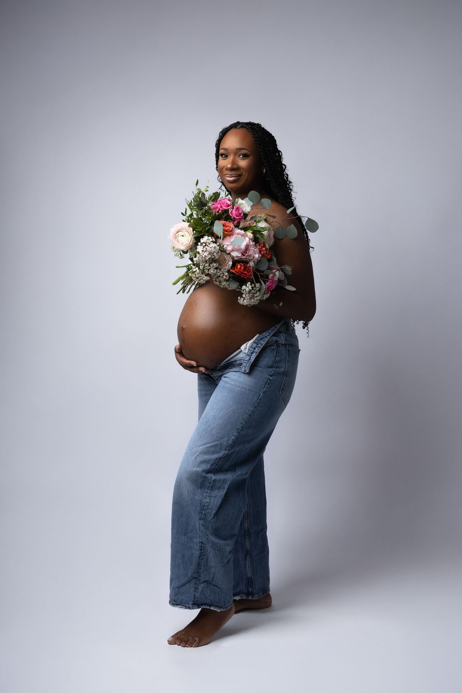 Smiling pregnant Black woman holding colorful flower bouquet against bare belly in studio portrait. photographe grossesse chavanoz lyon