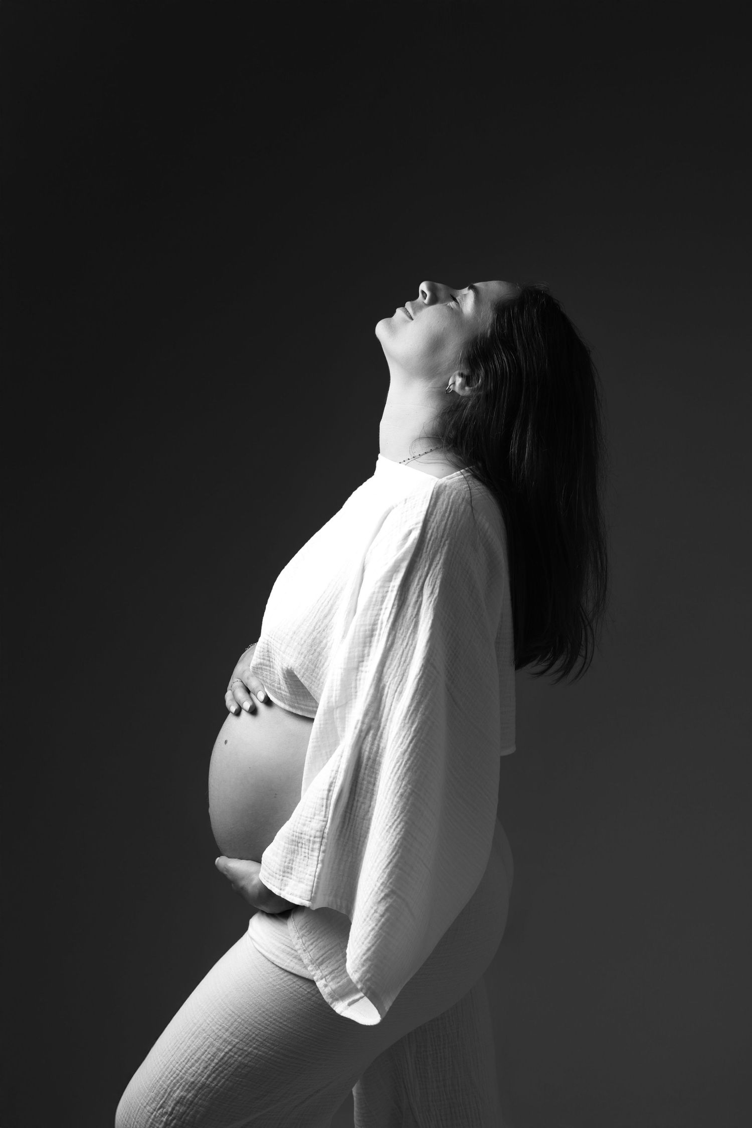 Black and white portrait of a pregnant woman in white top holding her baby bump, head tilted back on dark background.