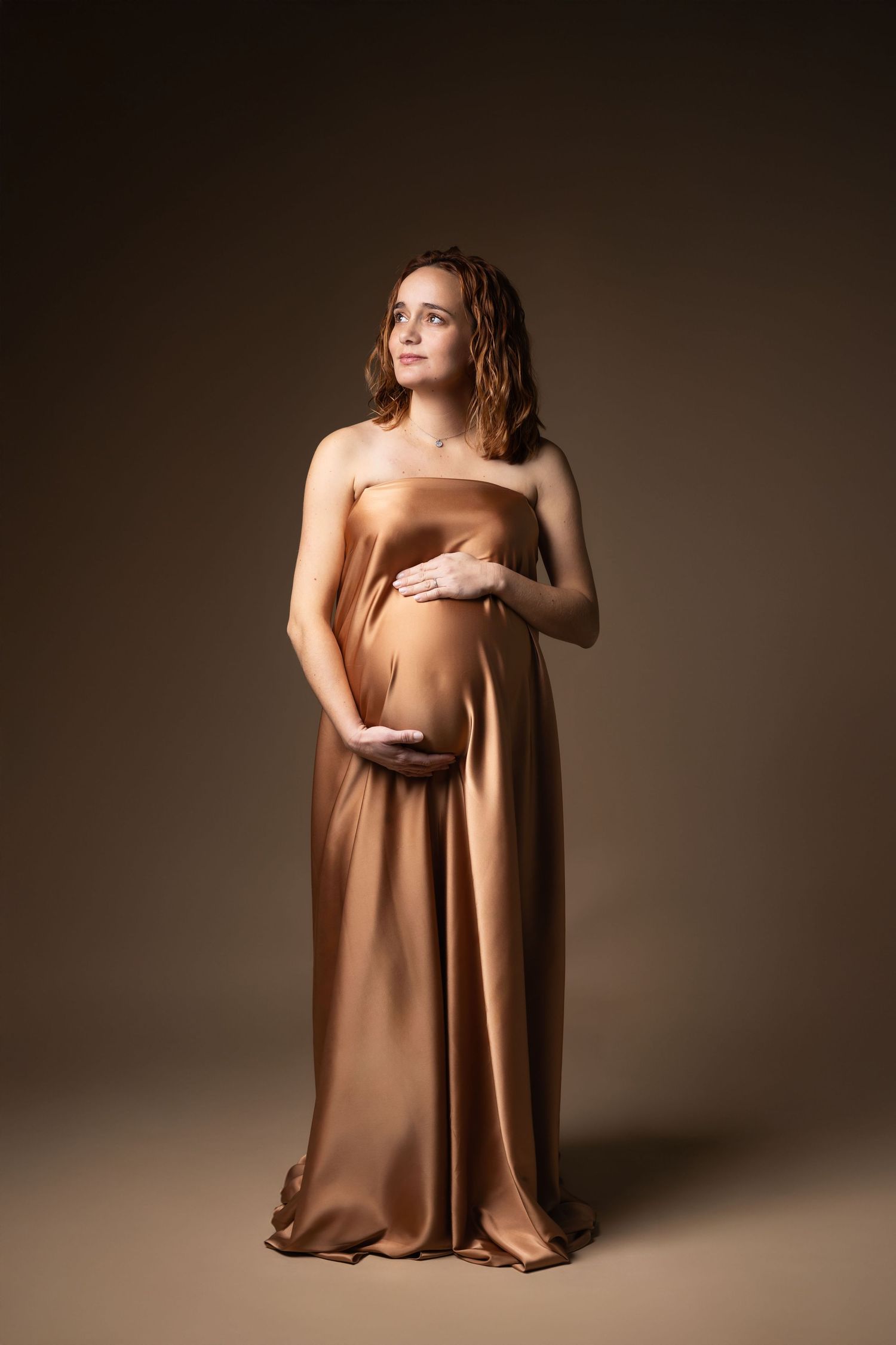Expectant mother in flowing gold satin wrap dress gently holding her pregnant belly, studio shot.