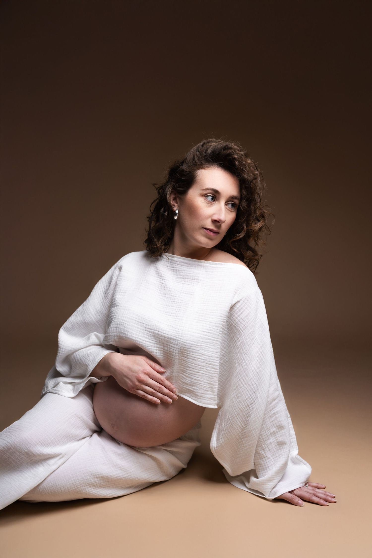 Curly-haired pregnant woman in white outfit sits on studio floor, gently holding her bare baby bump.