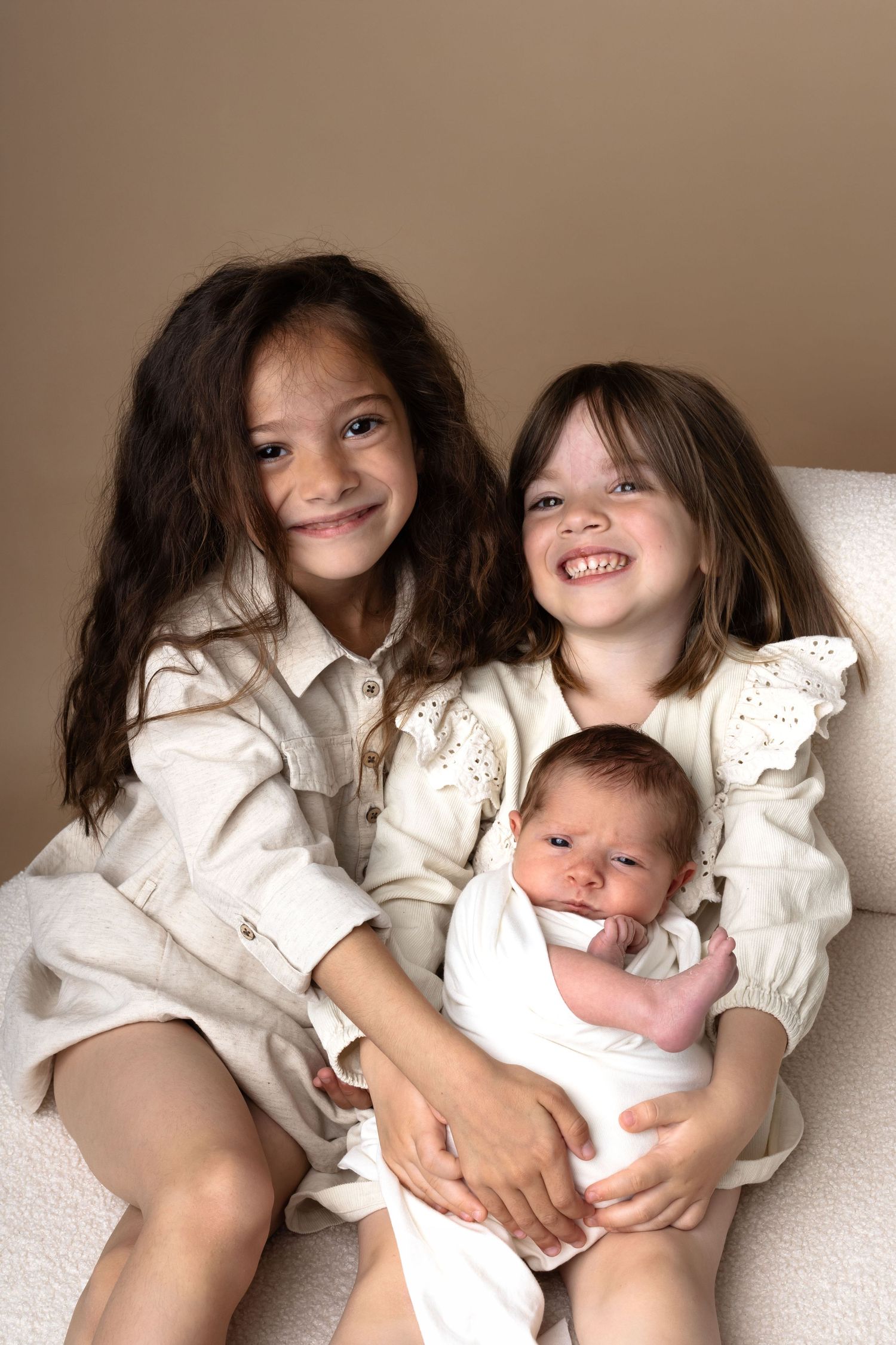 Two smiling girls in white dresses pose with a newborn baby in a cozy chair portrait.