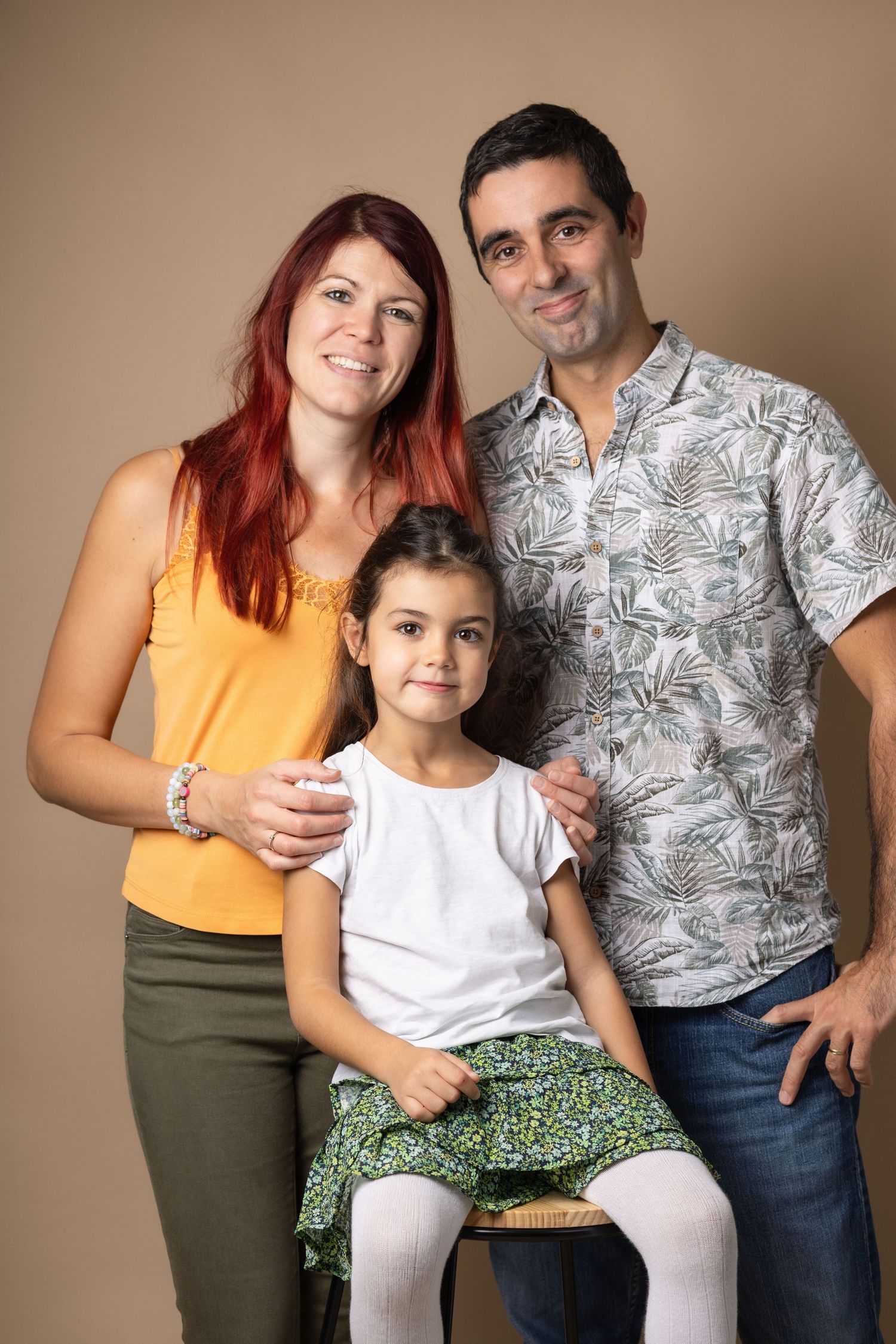A smiling family of three poses in a color studio portrait, with young girl seated between her parents.