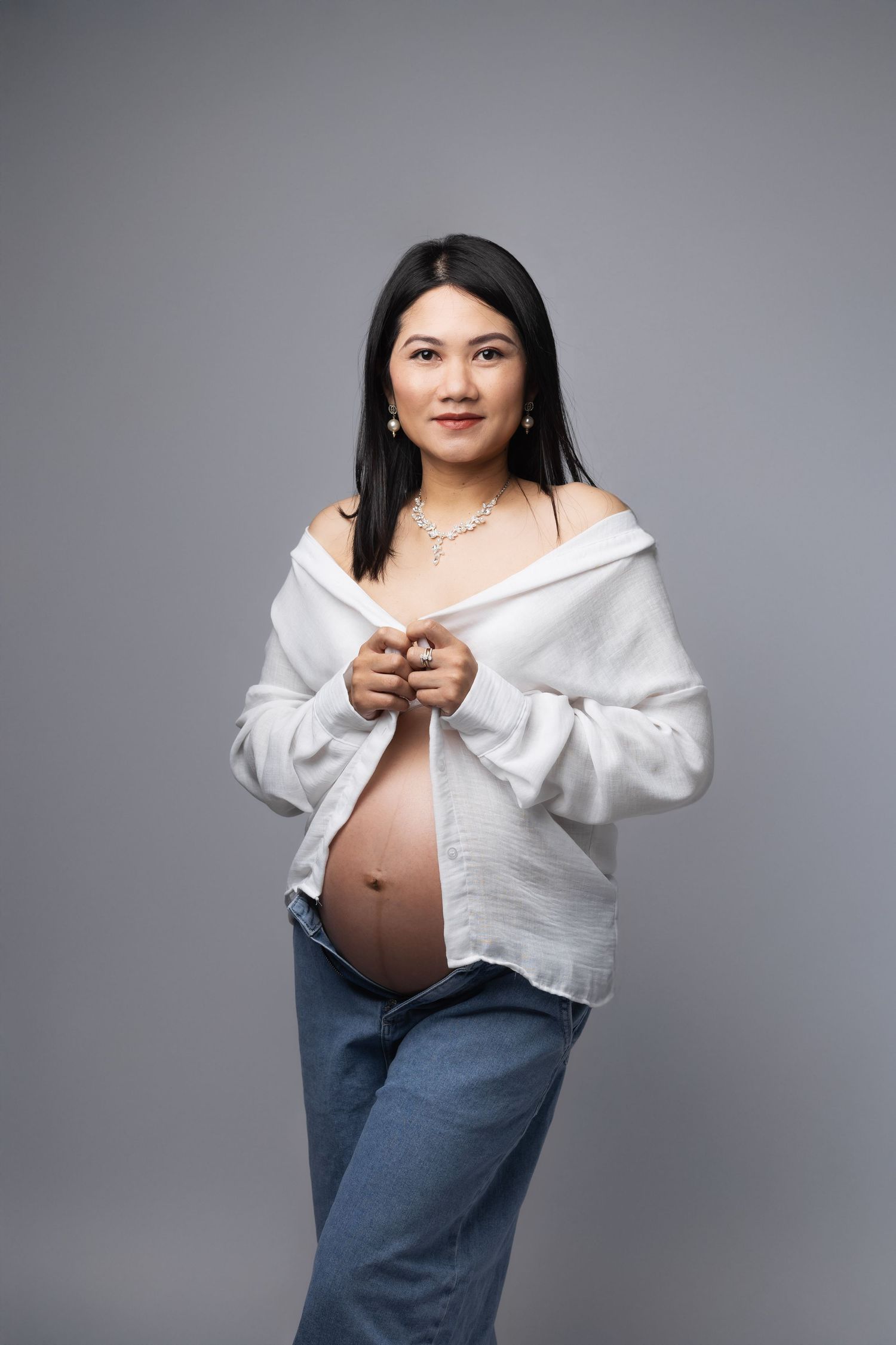 Smiling Asian pregnant woman in open white shirt and jeans reveals baby bump against grey studio background.