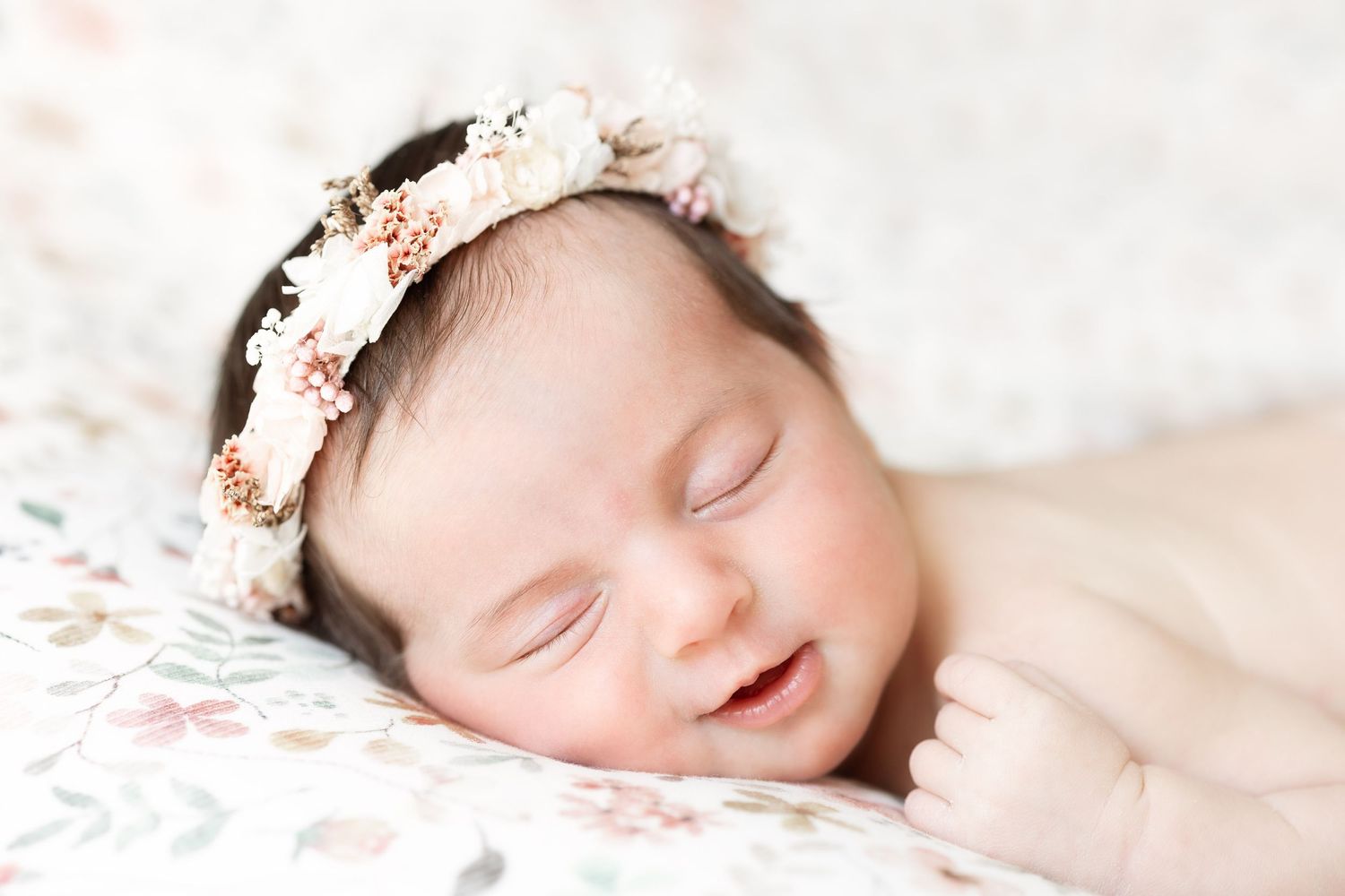 Smiling sleeping newborn baby girl wearing a delicate floral crown lying on a floral blanket.