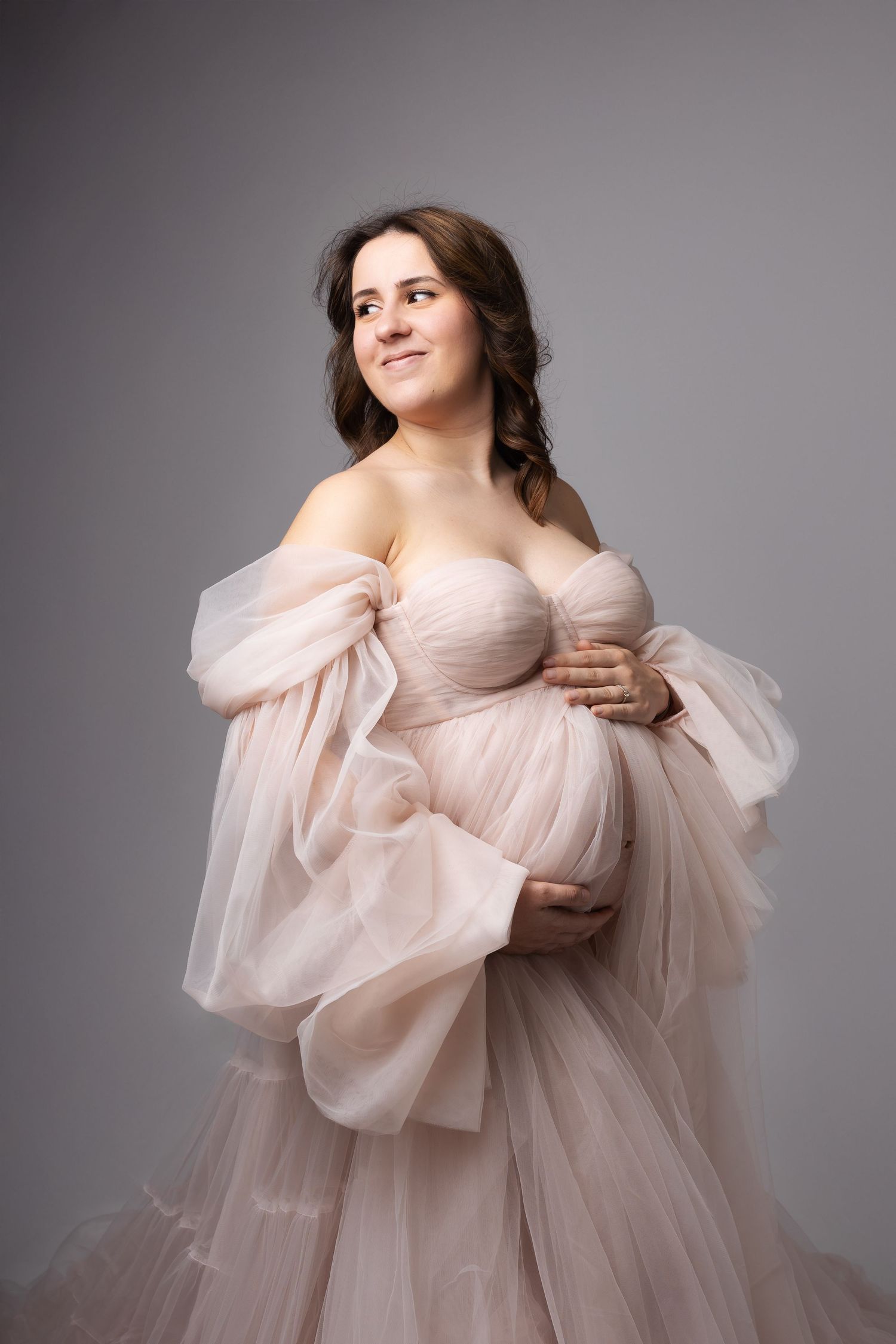 Pregnant woman in off-shoulder blush pink tulle gown cradling baby bump on grey background. photographe grossesse chavanoz lyon