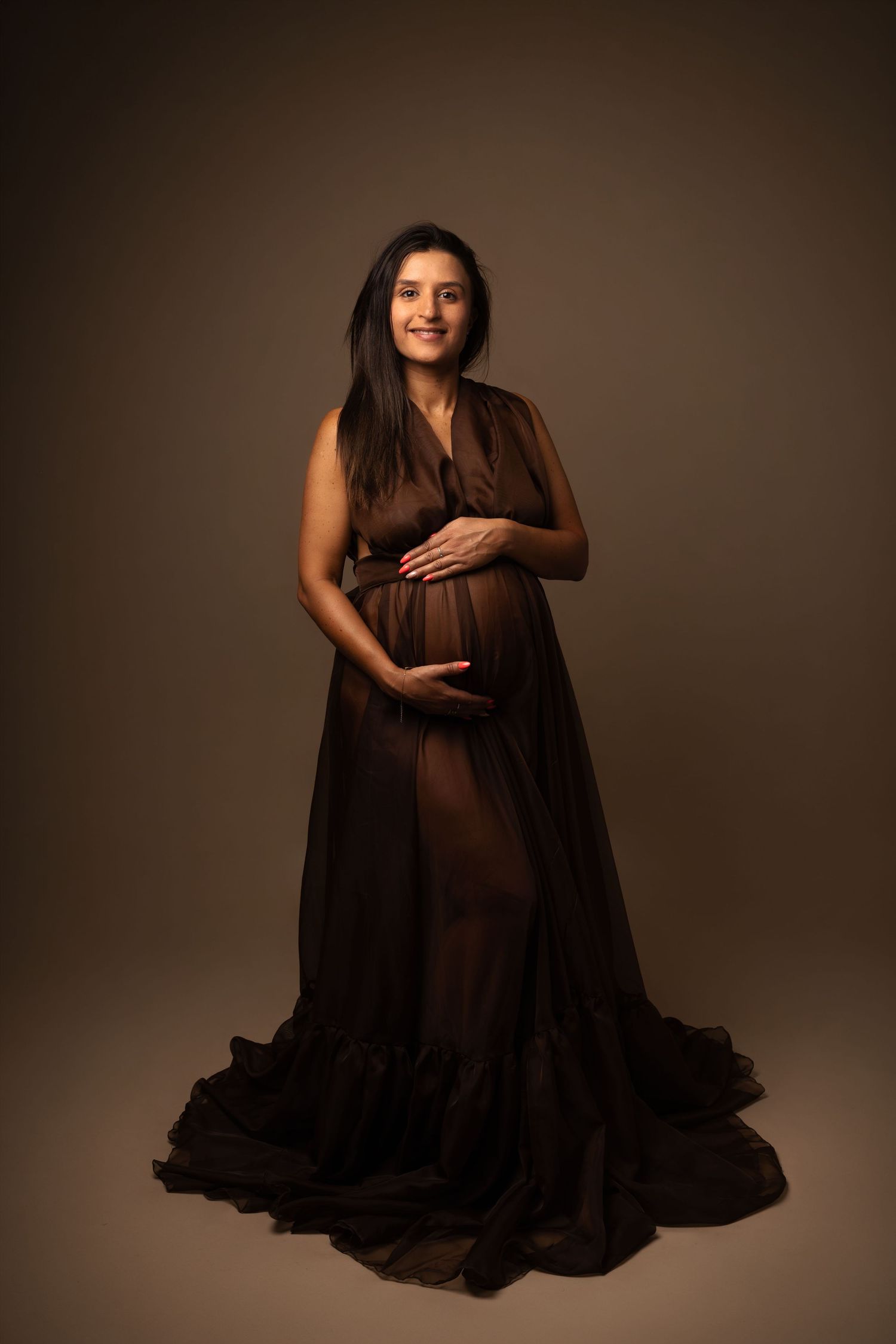 Pregnant woman in flowing brown chiffon gown cradling baby bump, elegant maternity studio portrait.