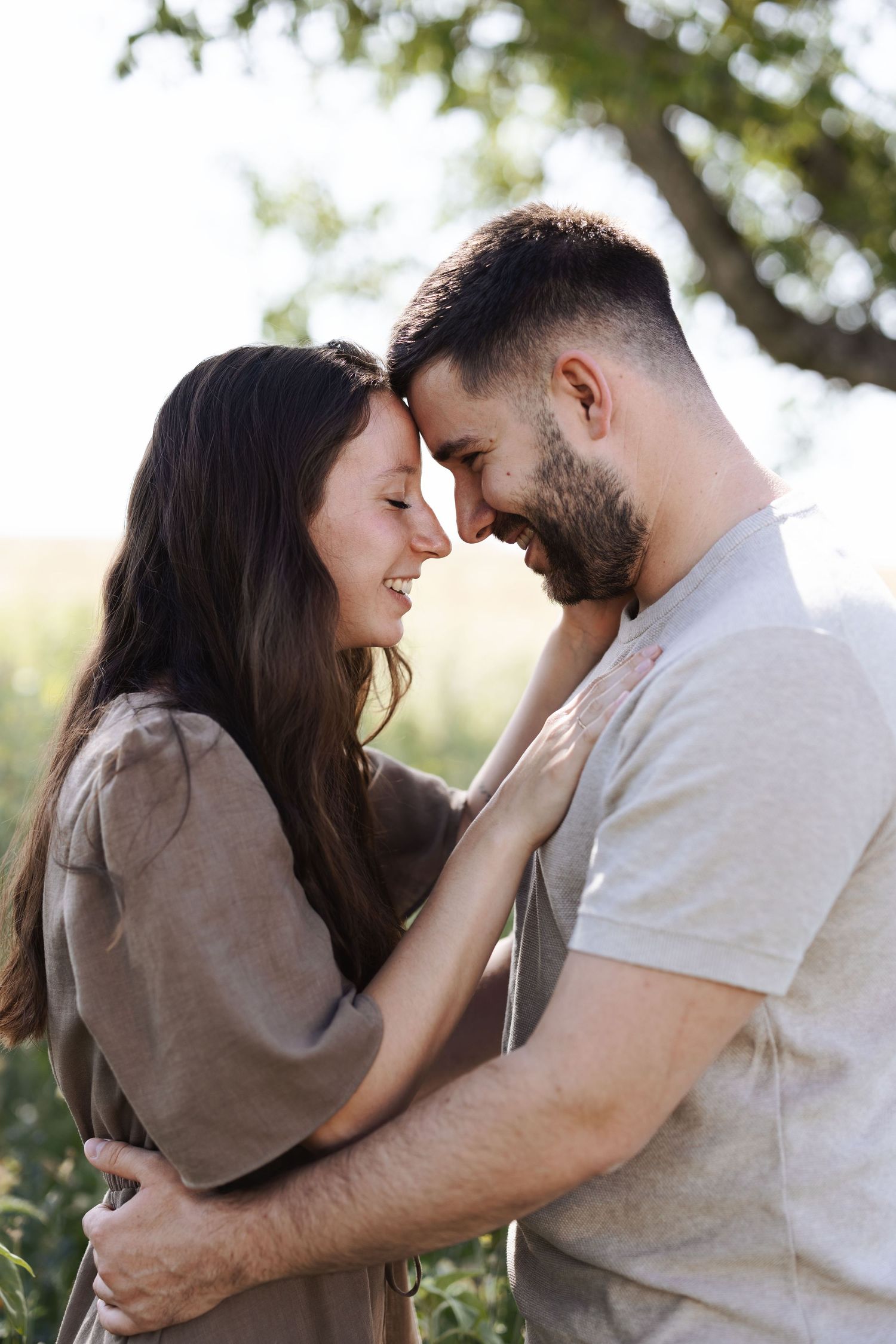 A happy couple embraces outdoors, touching foreheads and smiling, surrounded by lush green nature in warm sunlight.
