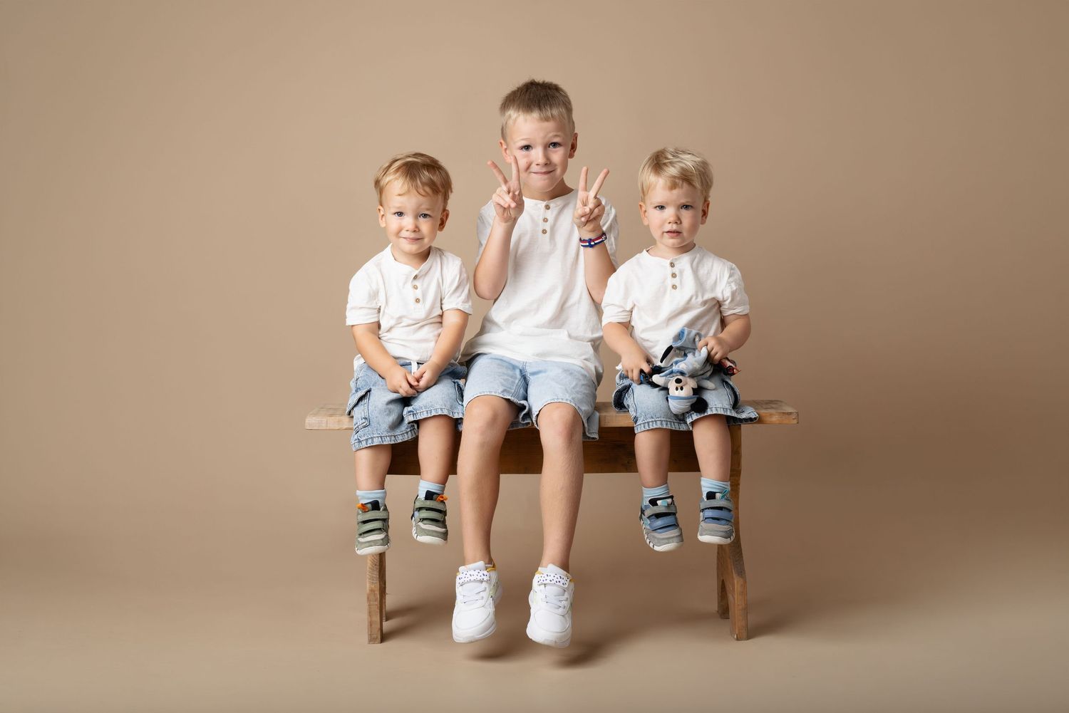 Three young boys in white shirts and blue shorts sit on a wooden bench, smiling and making hand gestures.