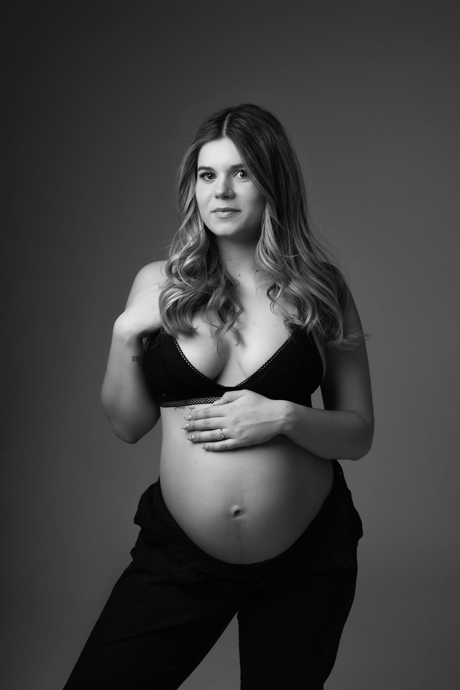 Black and white maternity portrait of pregnant woman in black bralette holding baby bump against gray background. photographe grossesse