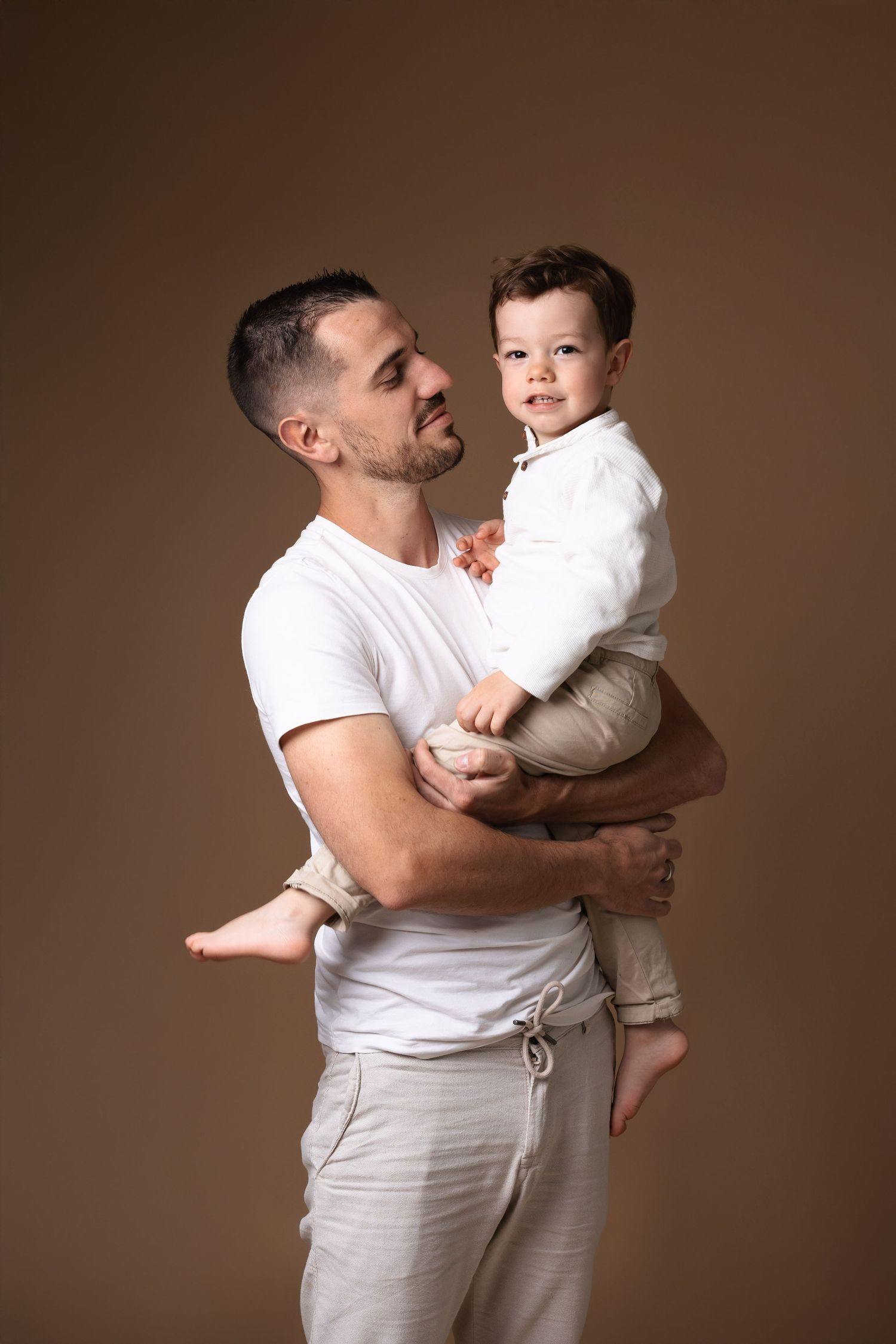 Father in white t-shirt holding his young toddler son on a warm brown background.