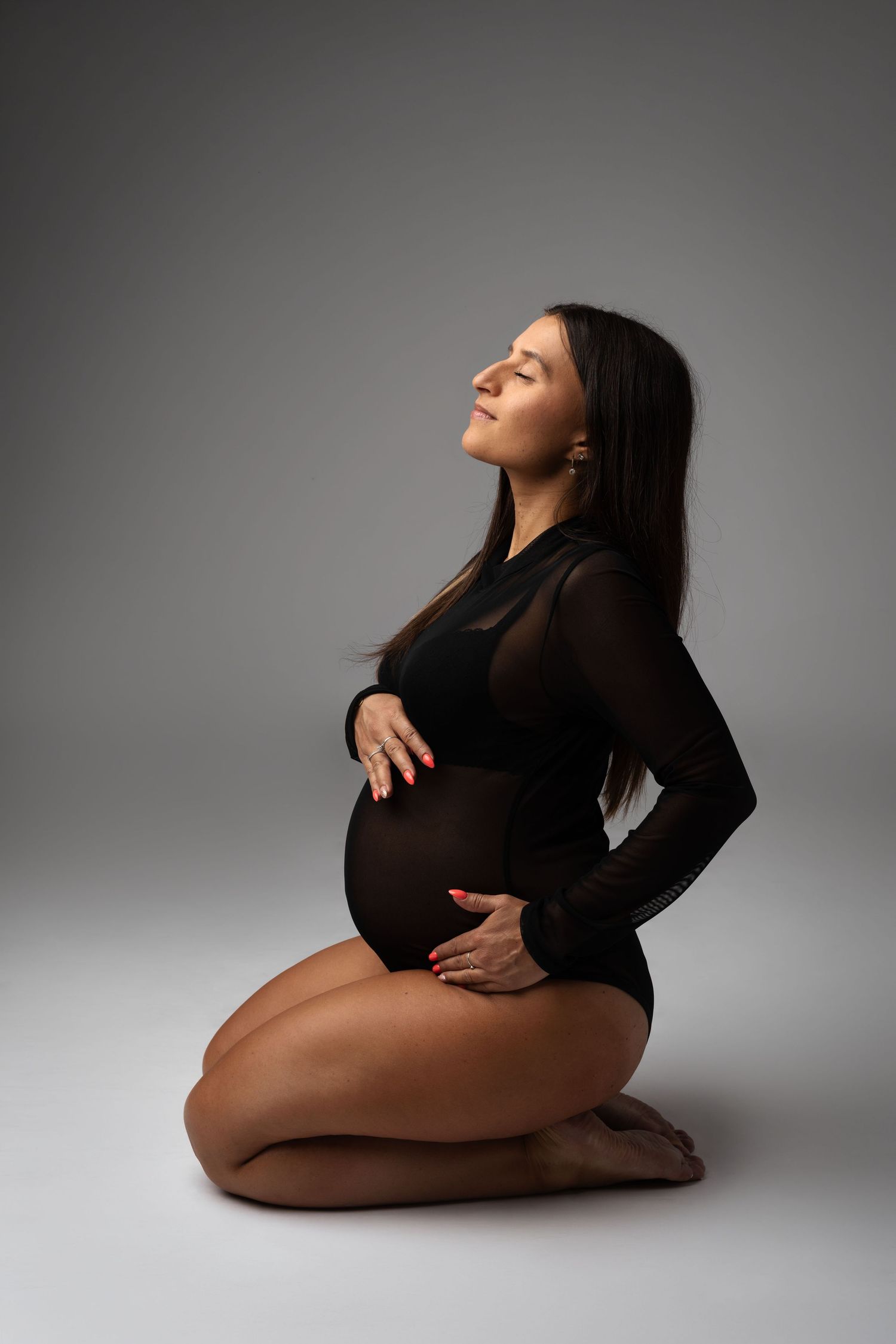 Pregnant woman in black bodysuit kneeling on floor, cradling baby bump with eyes closed on gray background.