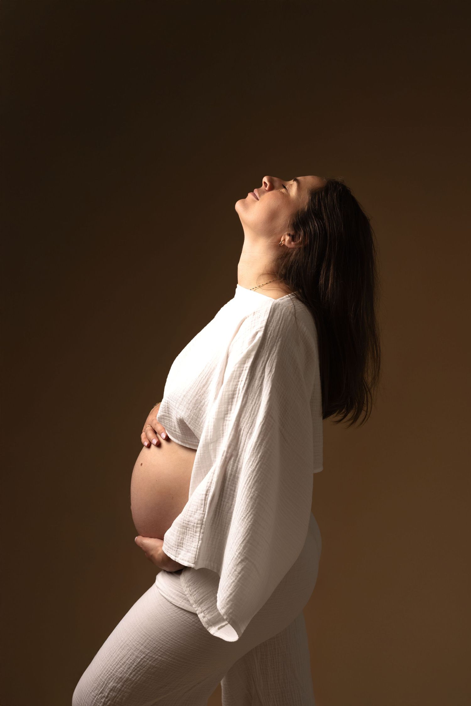 Pregnant woman in white outfit cradling her baby bump, head tilted back, against a warm brown background.