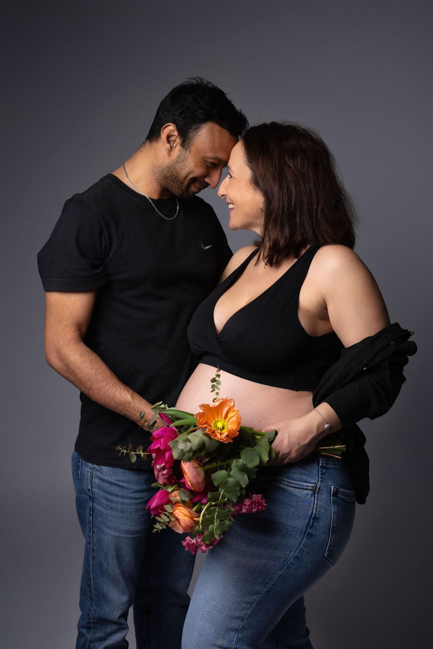Expecting couple in black outfits holding colorful flowers, foreheads touching in studio portrait. photographe grossesse chavanoz lyon