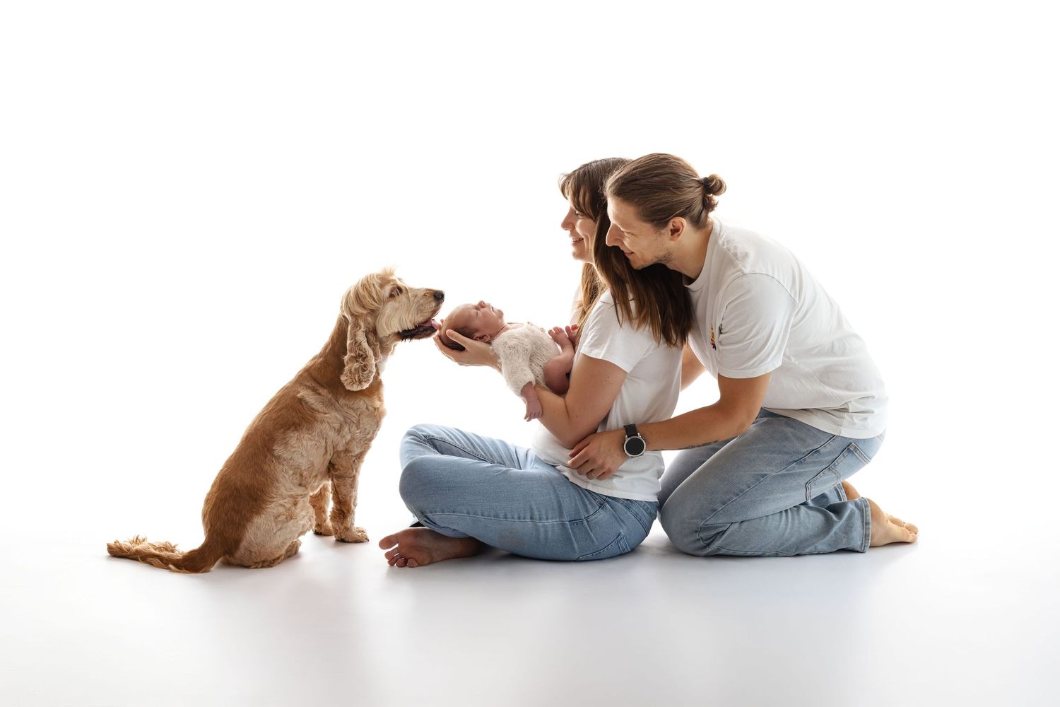 A couple sits on white floor with newborn baby while golden cocker spaniel dog interacts with the infant.