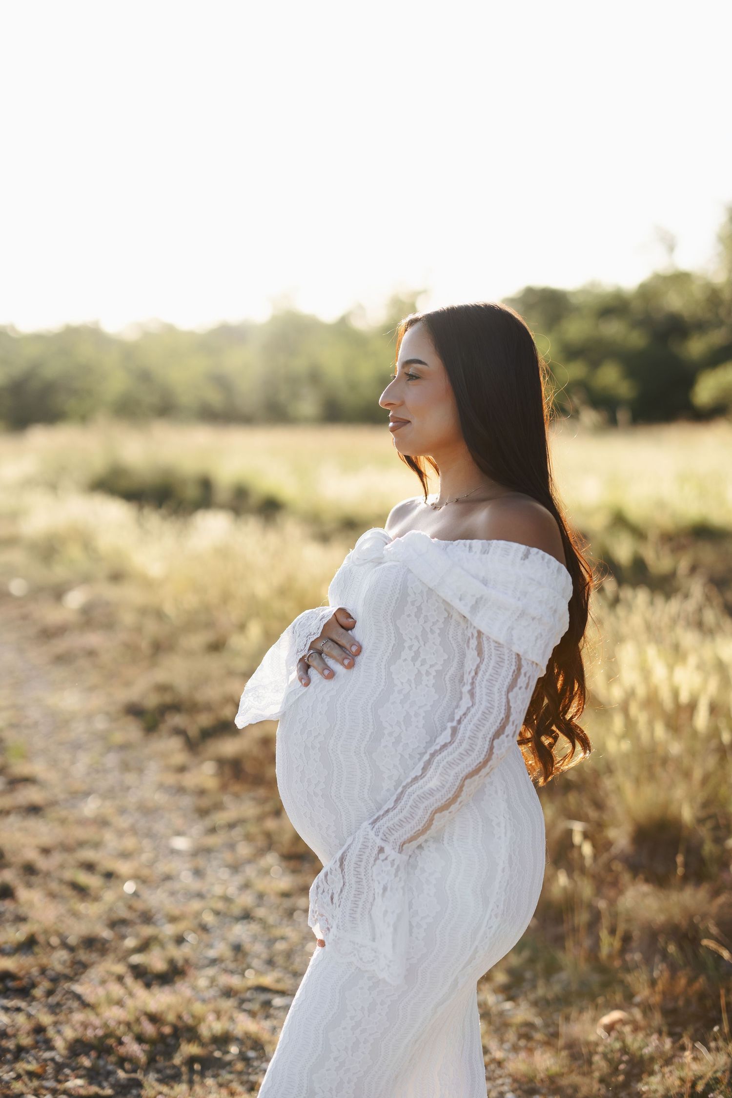 Pregnant woman in white lace off-shoulder dress cradling her baby bump in a sunlit field.