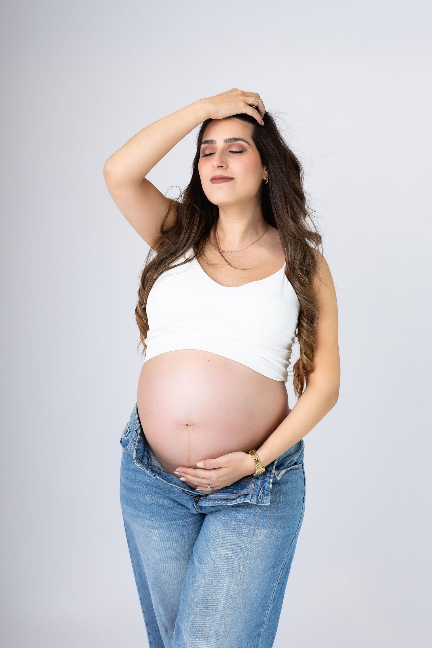 Pregnant woman in white crop top and jeans holding her bare bump with hand raised to her head. photographe grossesse chavanoz lyon