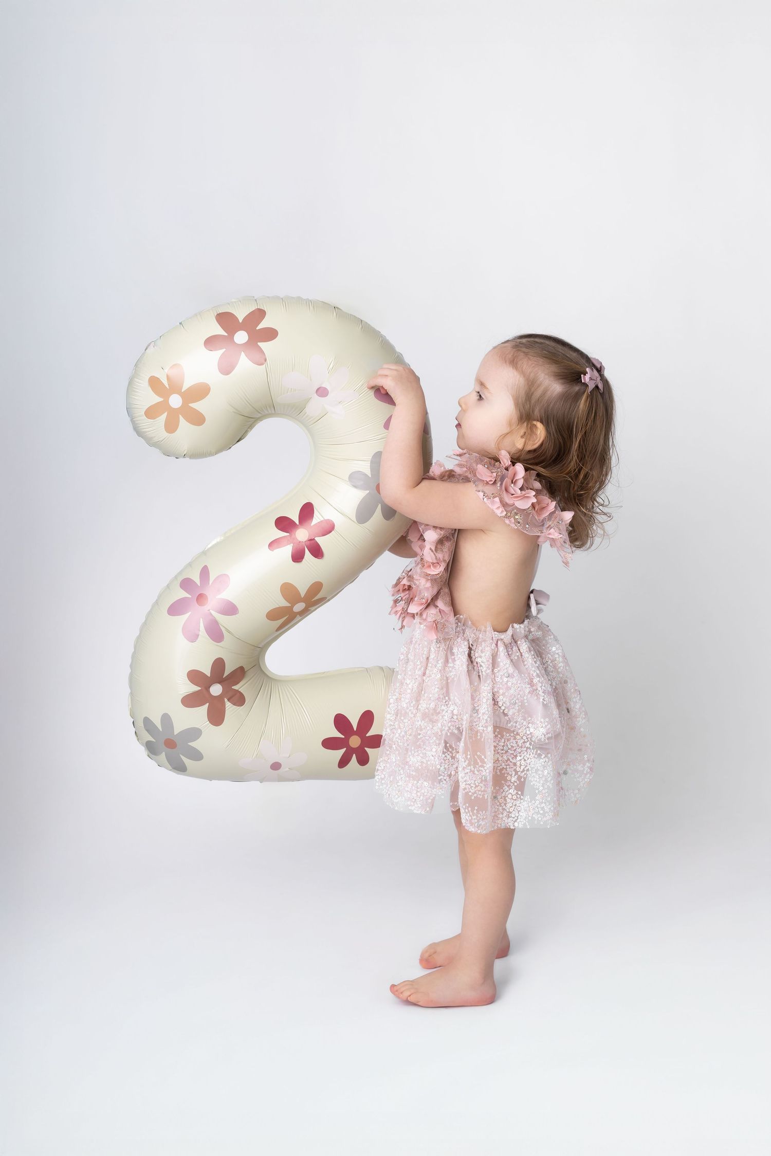 Toddler girl in pink floral dress stands holding a large number 2 balloon with flower prints.