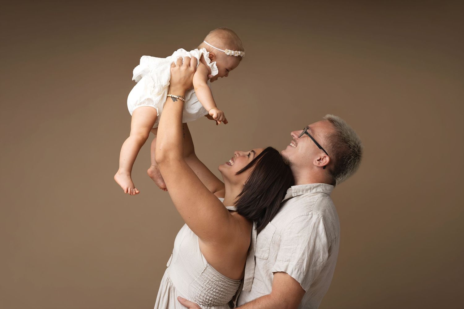 Parents joyfully lifting their smiling baby girl in white dress during professional family portrait studio session.