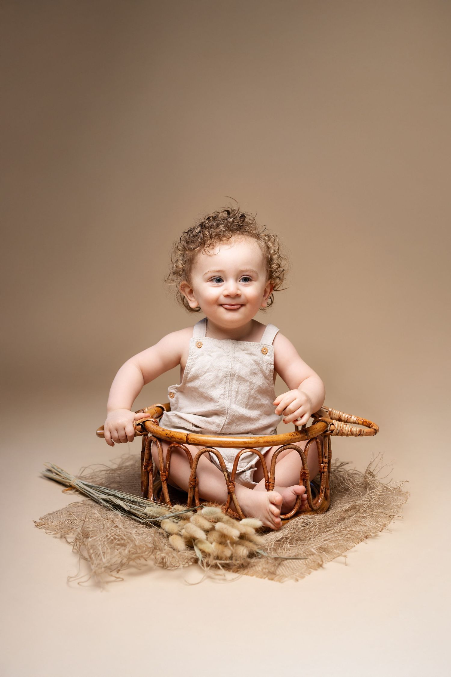 Adorable curly-haired baby sitting in wicker basket smiling on beige background in studio.