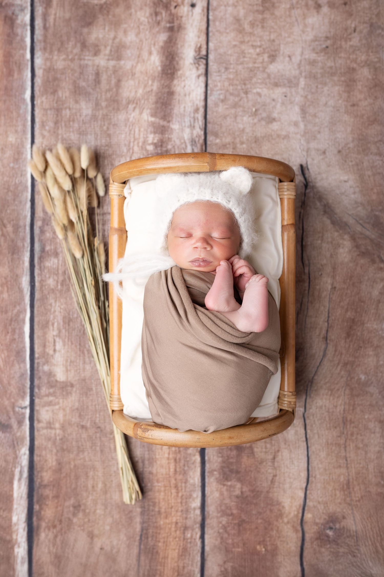Newborn baby swaddled in brown wrap sleeping in a small bamboo crib on a rustic wooden floor. séance naissance chavanoz lyon