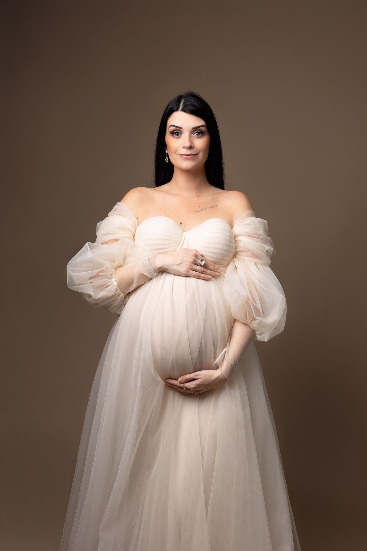 Elegant pregnant woman in cream off-shoulder tulle gown cradling baby bump against warm brown background.