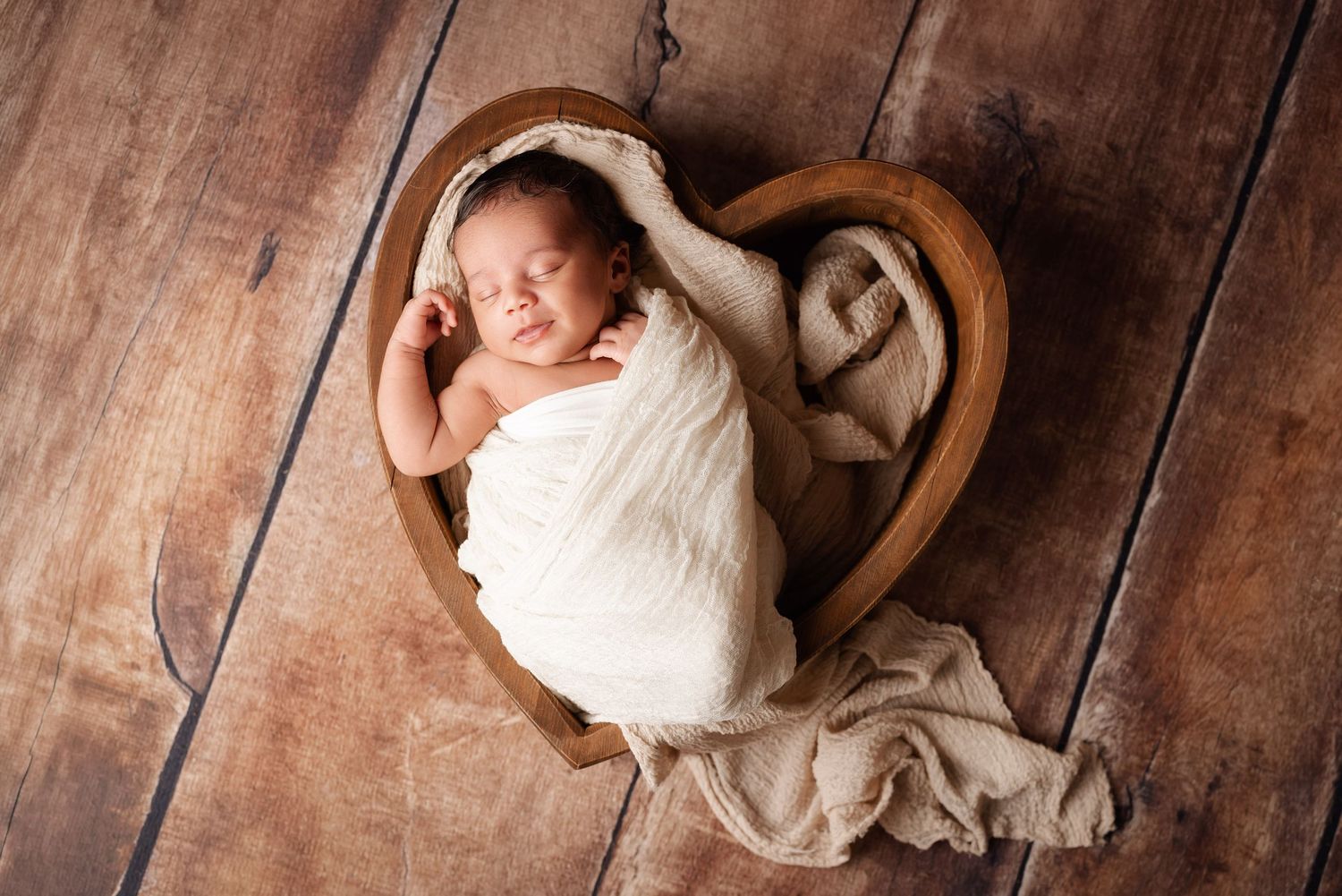 Newborn baby swaddled in white wrap sleeping peacefully in a wooden heart-shaped bowl on rustic wood floor.