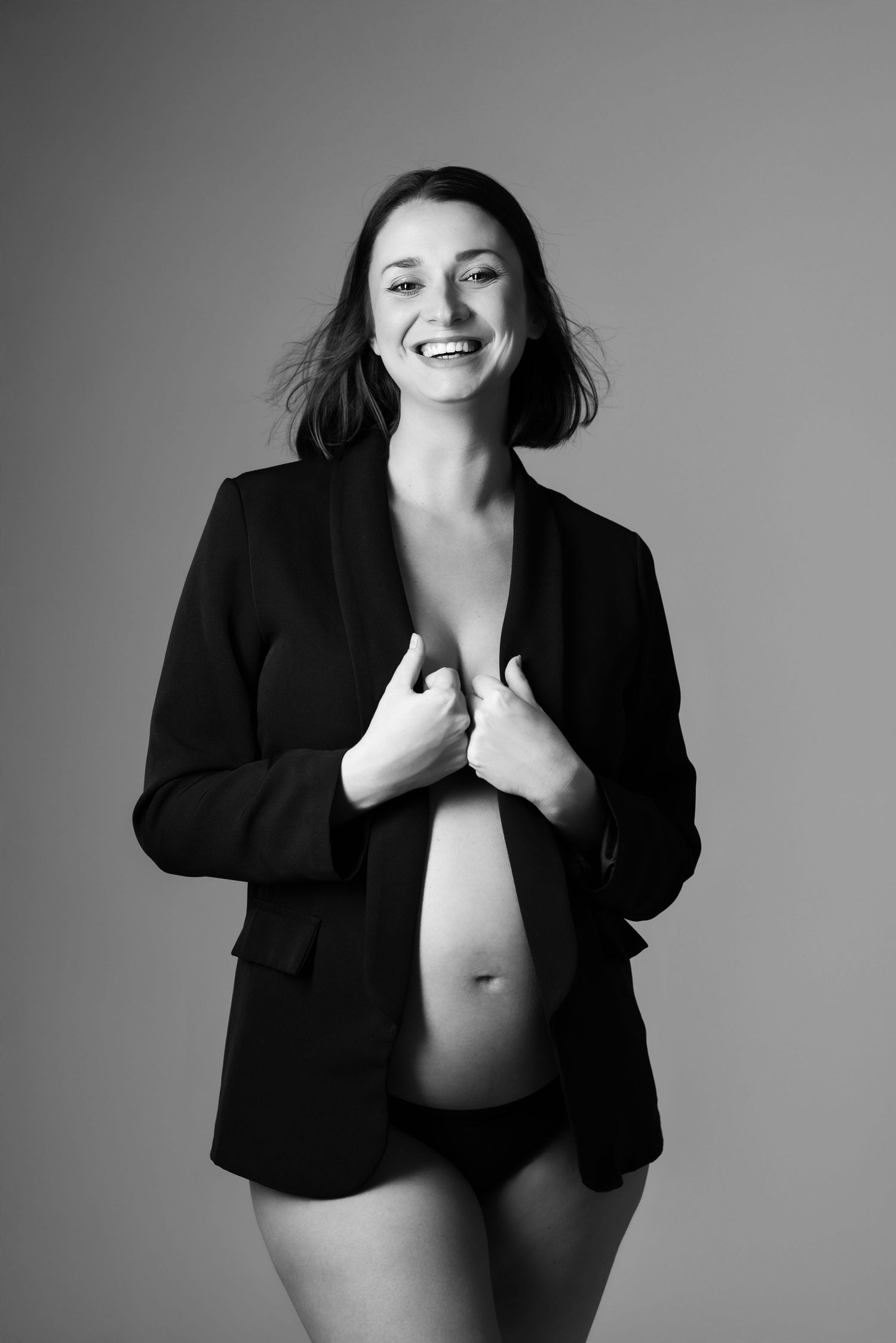 Black and white photo of laughing pregnant woman in blazer and underwear holding her baby bump.
