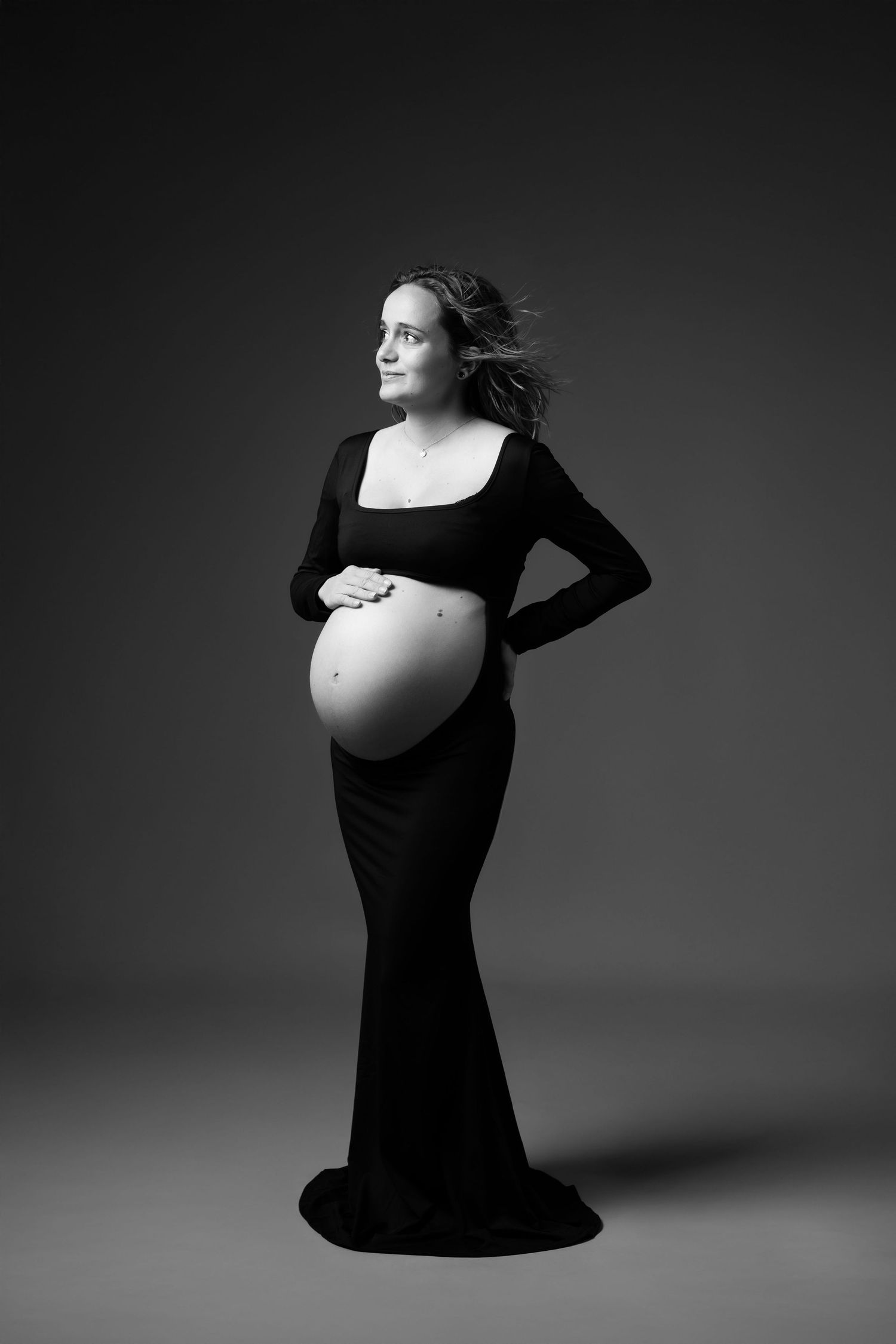 Black and white full-length studio portrait of pregnant woman in black gown with bare belly.
