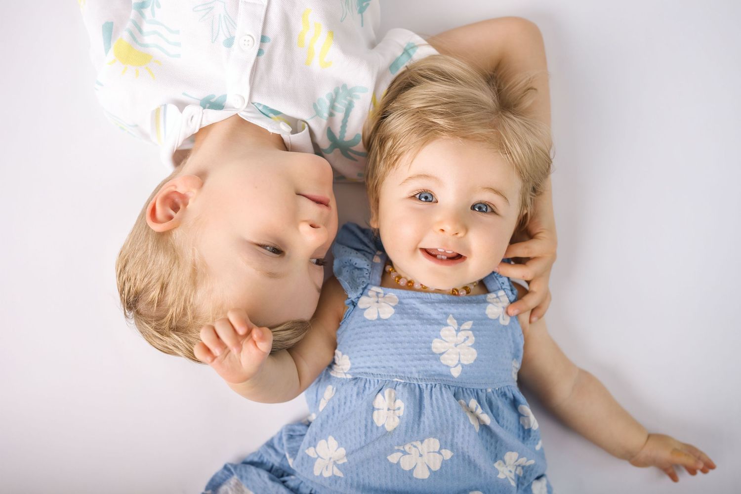 Two young blonde siblings laying together on a white floor, sharing a joyful moment.
