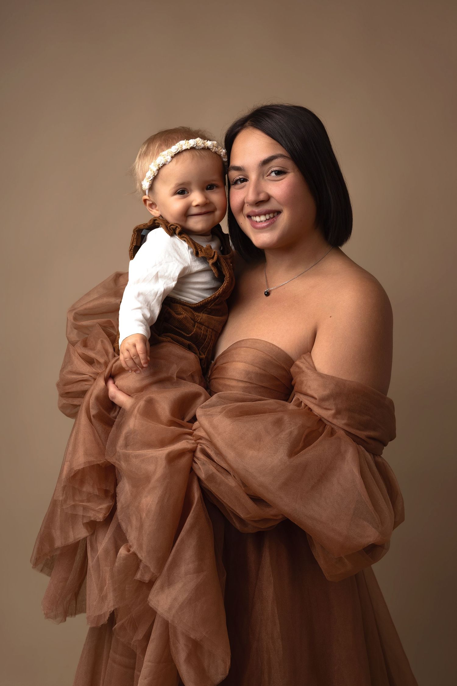 A young woman in a brown tulle gown holds a smiling baby girl in a brown outfit against a neutral backdrop.