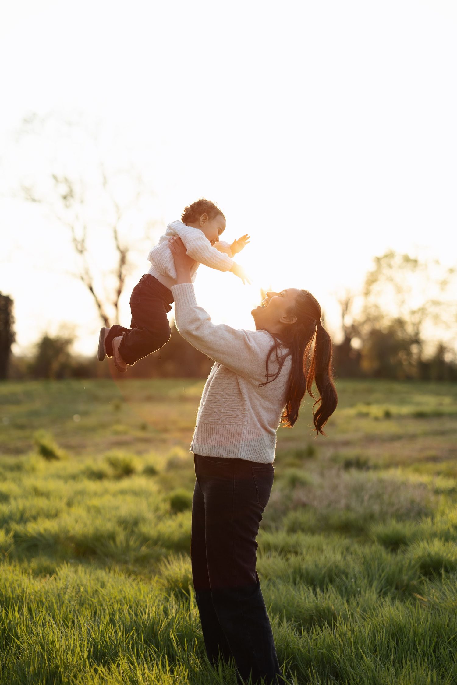 A mother joyfully lifts her baby in the air in a sunlit green field at golden hour.