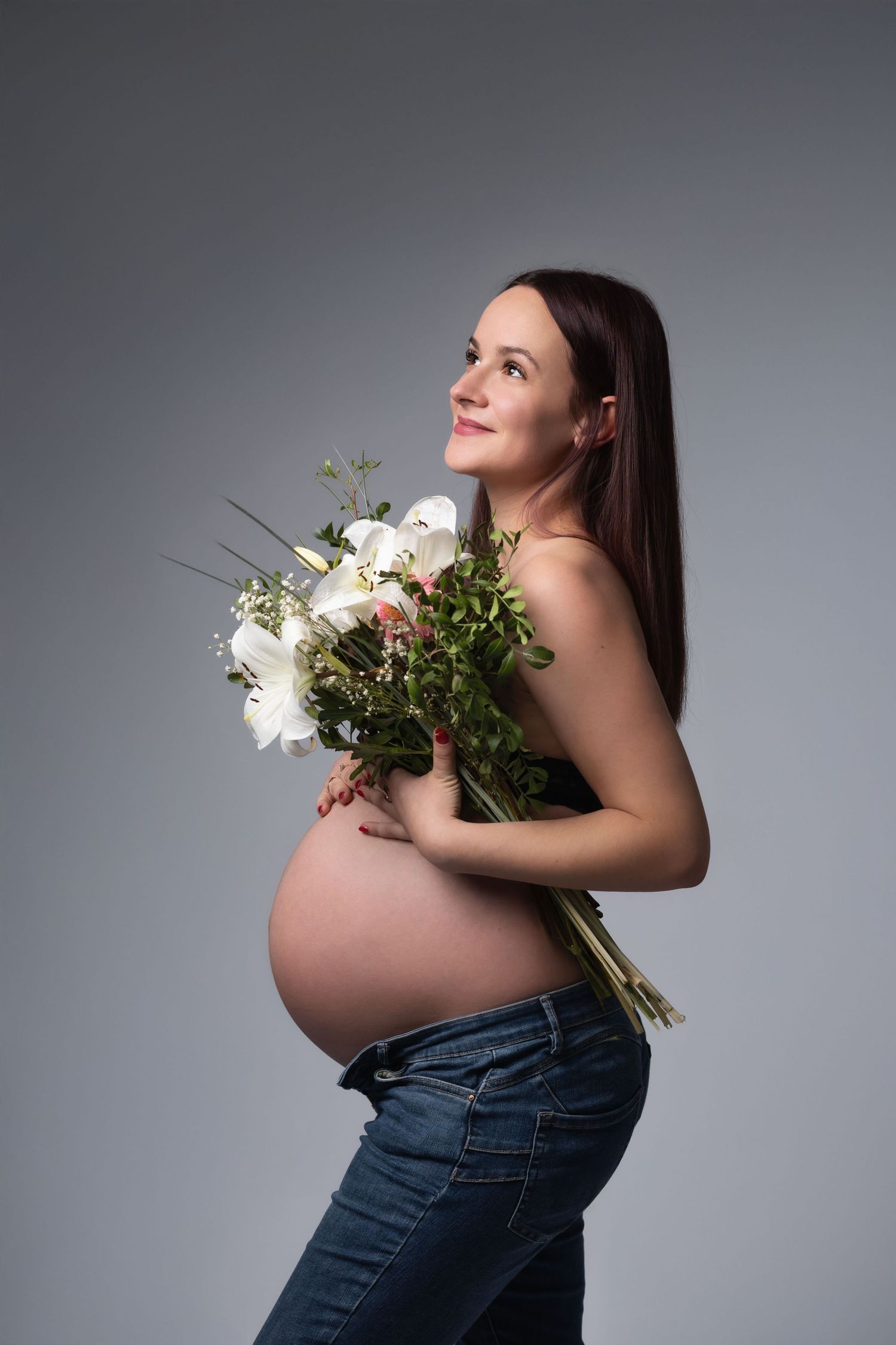 Pregnant woman holding a bouquet of white flowers, wearing jeans, smiling in a studio.