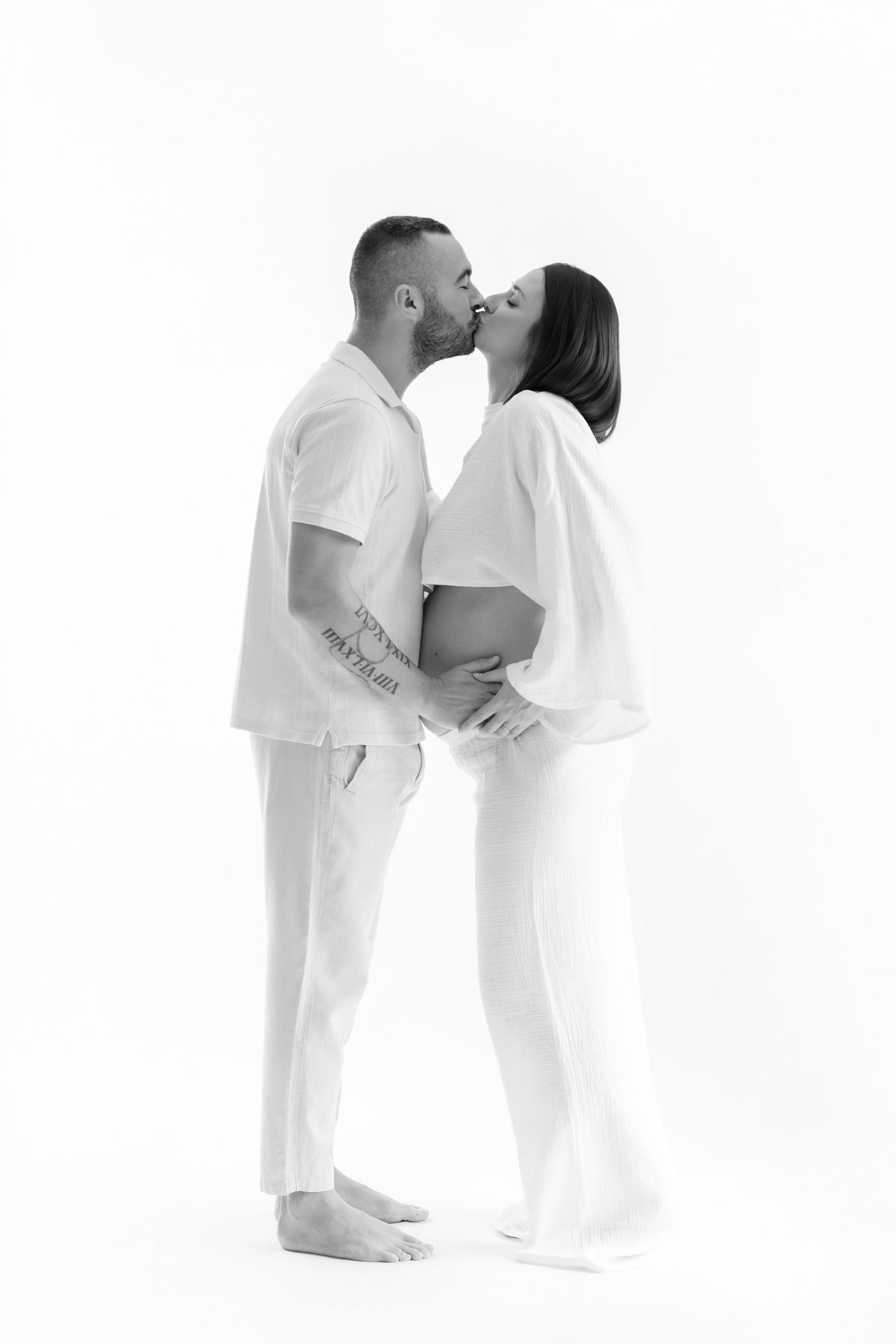 Black and white photo of a couple kissing while cradling a pregnant belly in a studio setting.