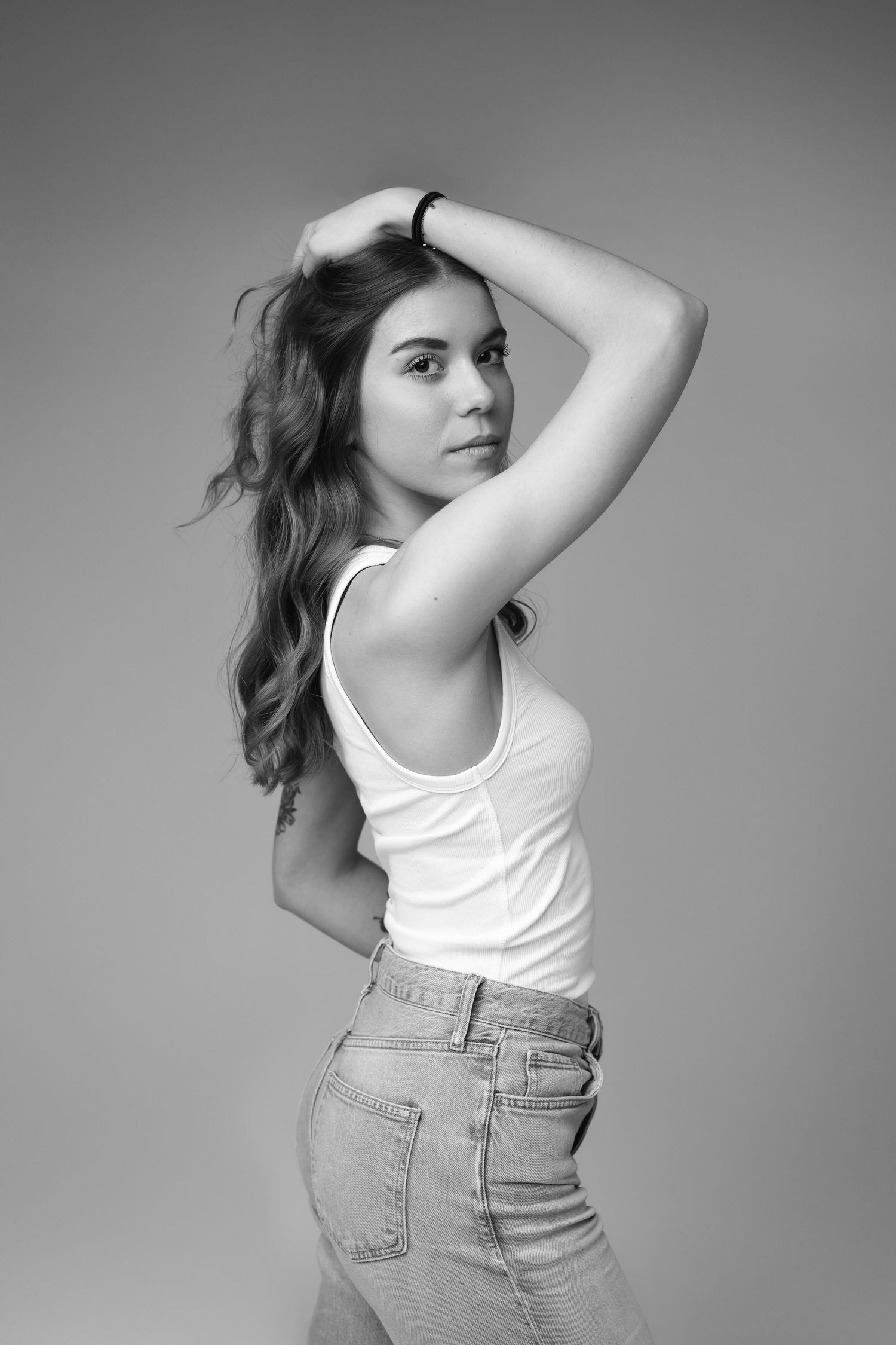Black and white portrait of young woman in white tank top posing with hand in hair.