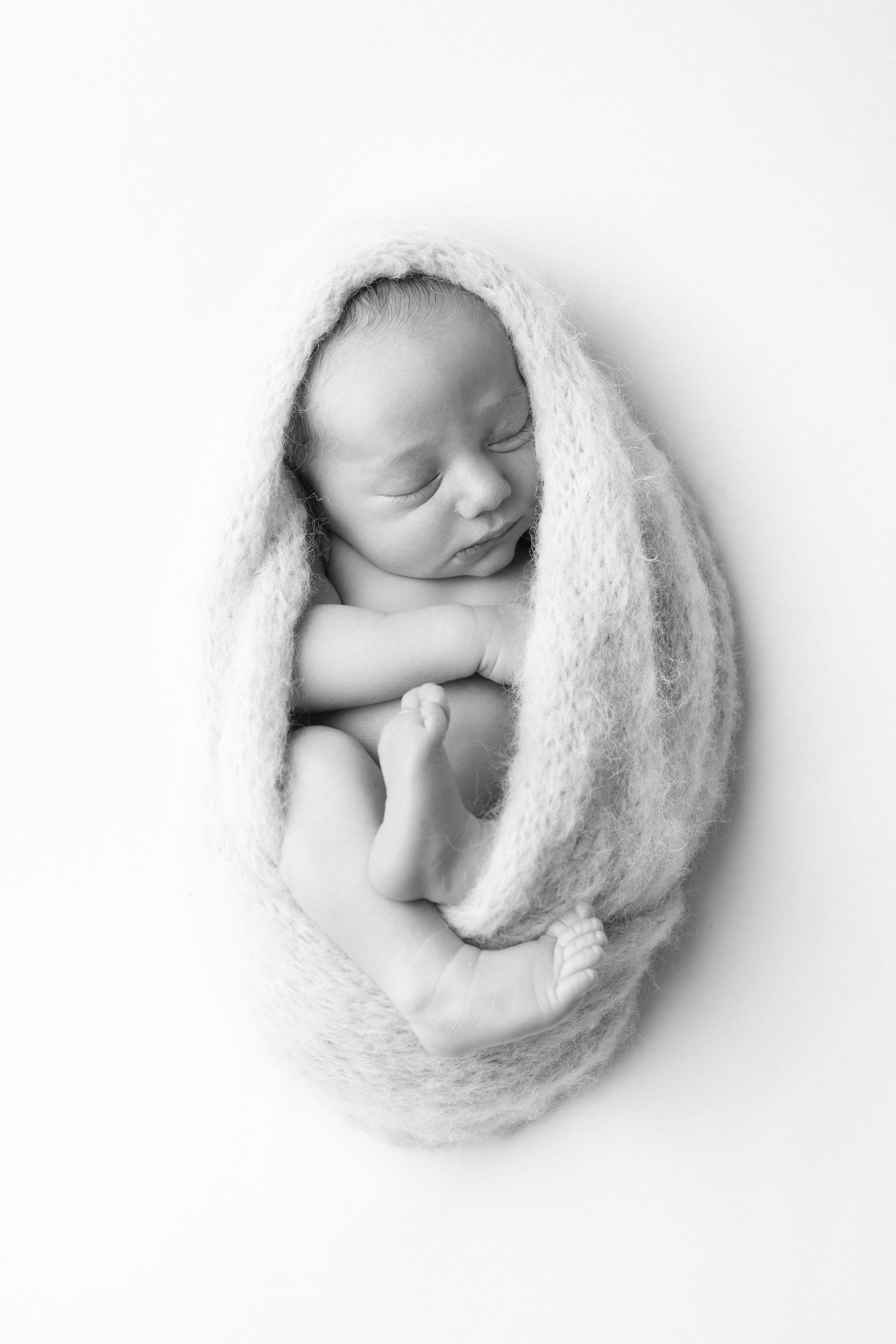 Black and white photo of a sleeping newborn baby wrapped snugly in a soft knit blanket on white background.
