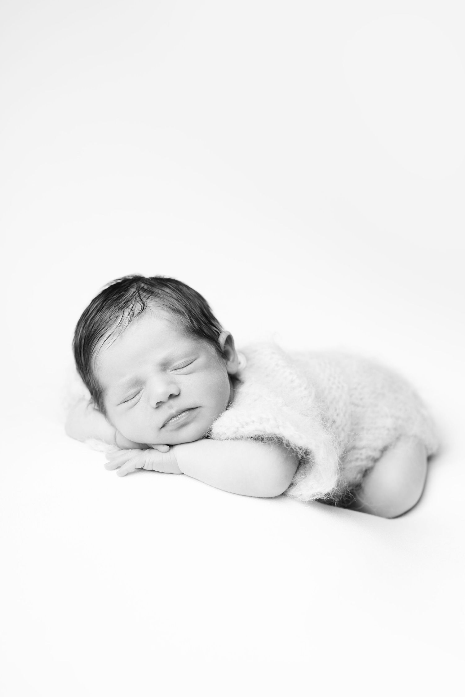 Black and white photo of a peaceful sleeping newborn baby wearing a soft knit romper, resting chin on hands.