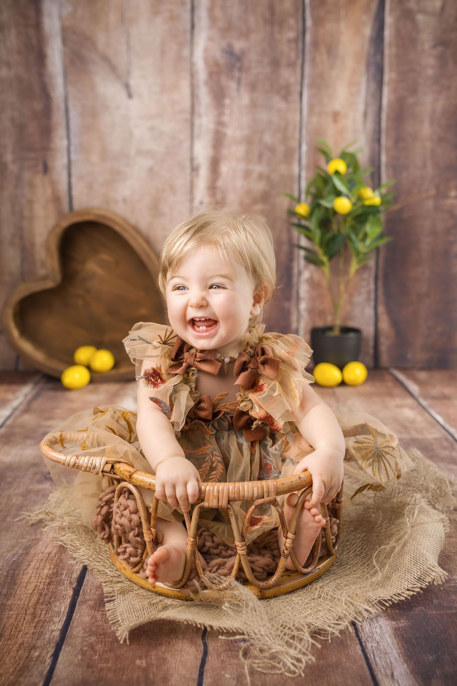 Laughing baby girl in a brown ruffled dress sits in a wicker basket with rustic wood backdrop.