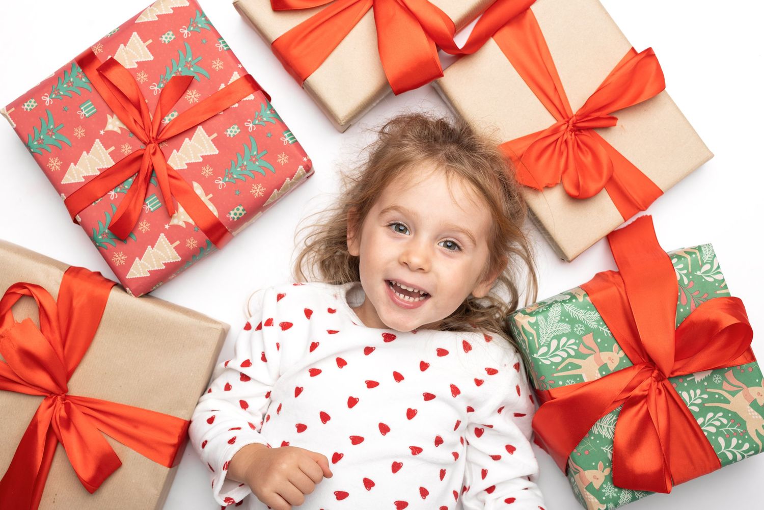 Happy young girl lying among colorful Christmas gift boxes with red ribbons, wearing a white heart-print shirt.