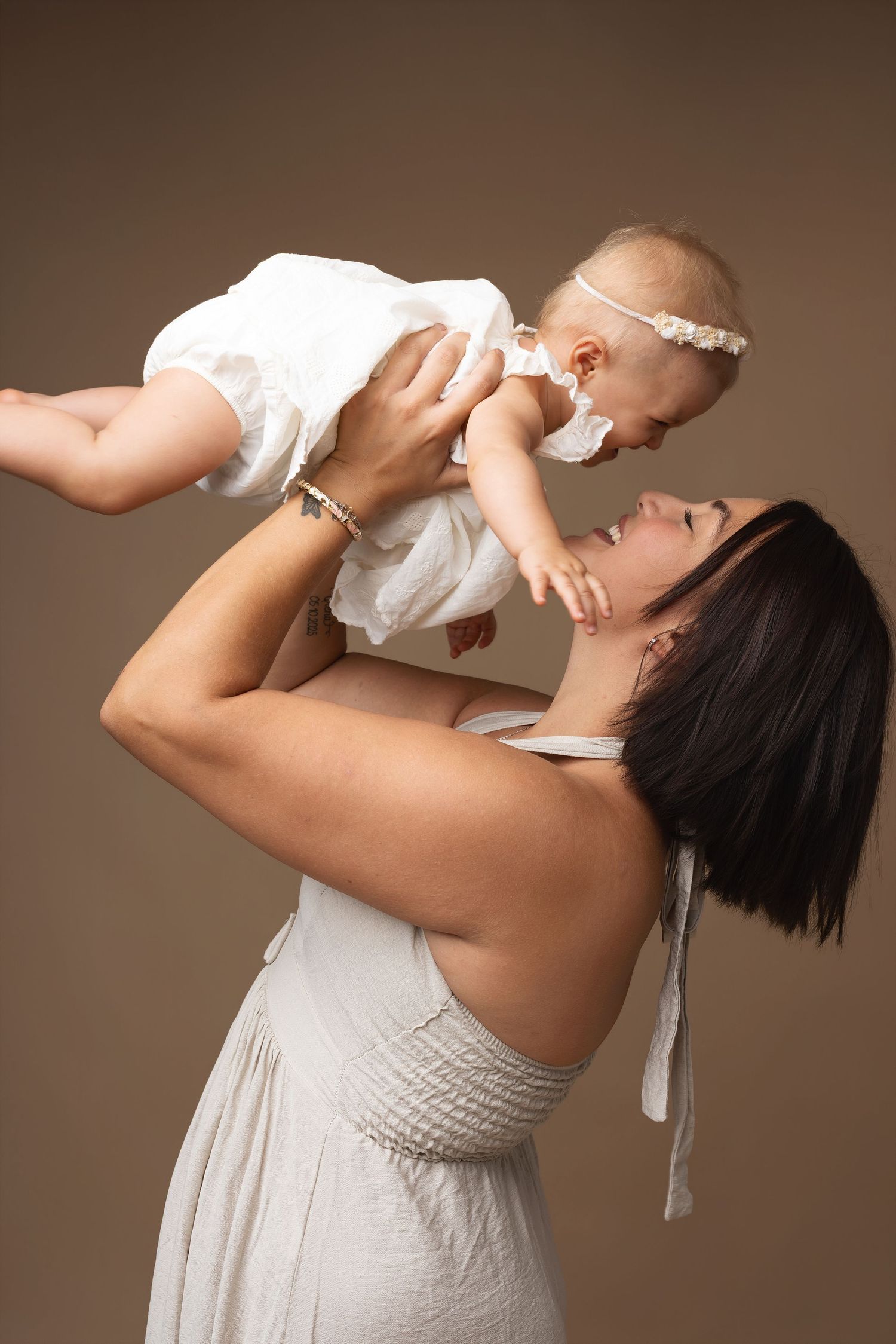A joyful mother in a white dress lifts her smiling baby girl wearing a white outfit and headband against a brown background.
