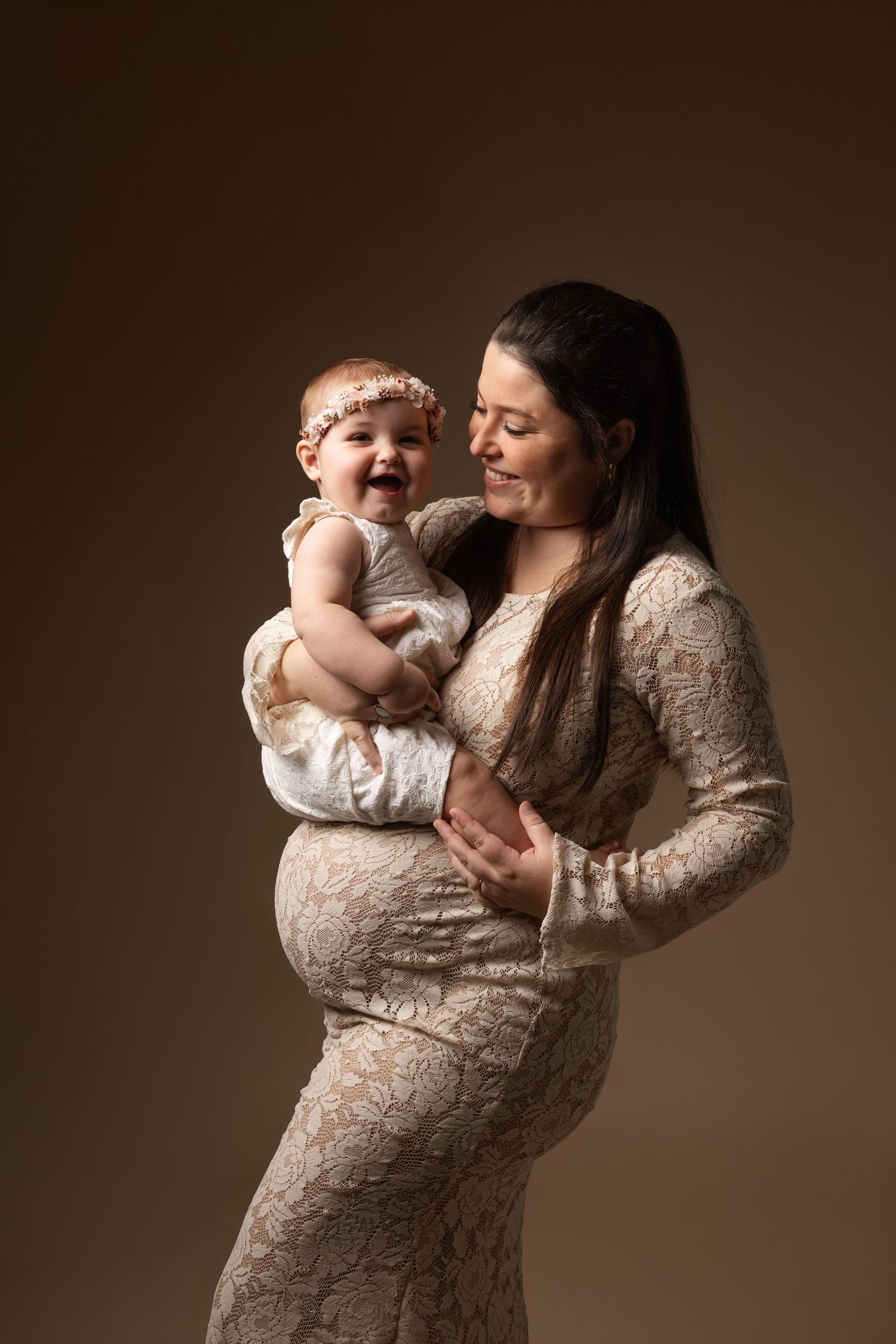 Pregnant woman in lace dress holds laughing baby girl with floral headband against brown studio background.