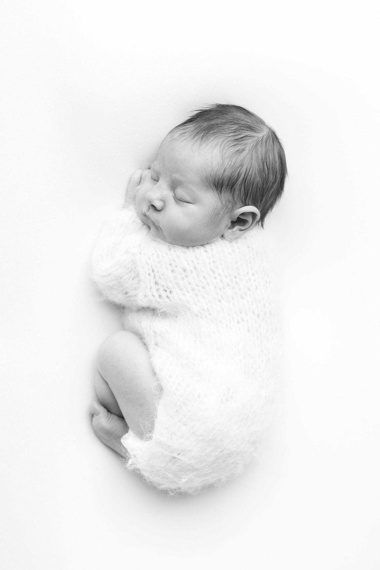 Black and white photo of a peaceful sleeping newborn baby wrapped in a soft white knit blanket.