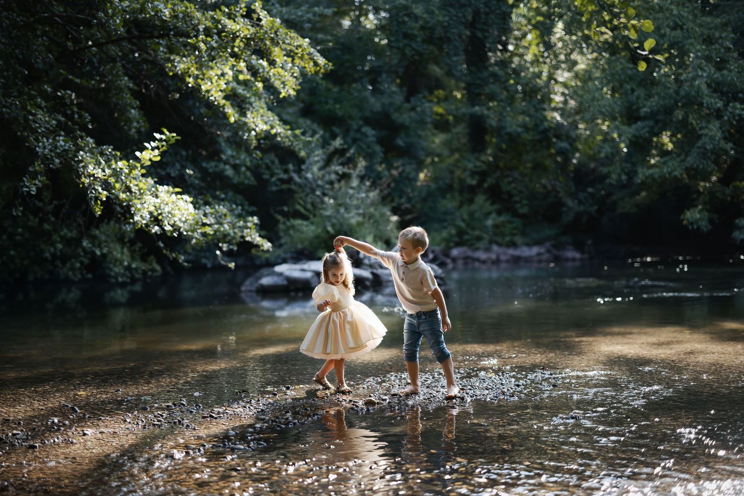 Two children playing in a shallow forest creek, girl in white dress and boy in casual clothes, surrounded by lush green trees.