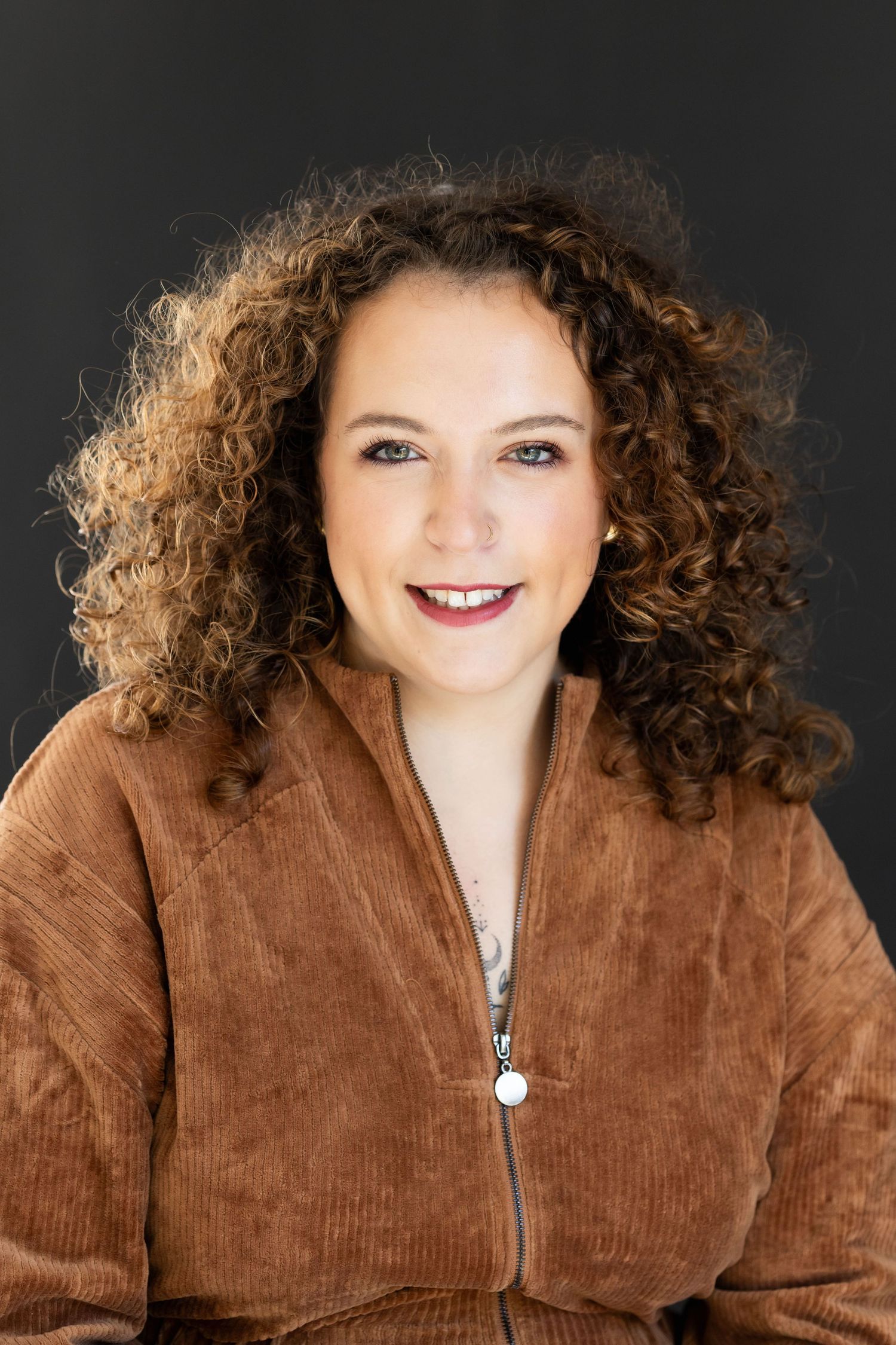 Portrait of cheerful woman with curly auburn hair wearing brown corduroy jacket on dark background.