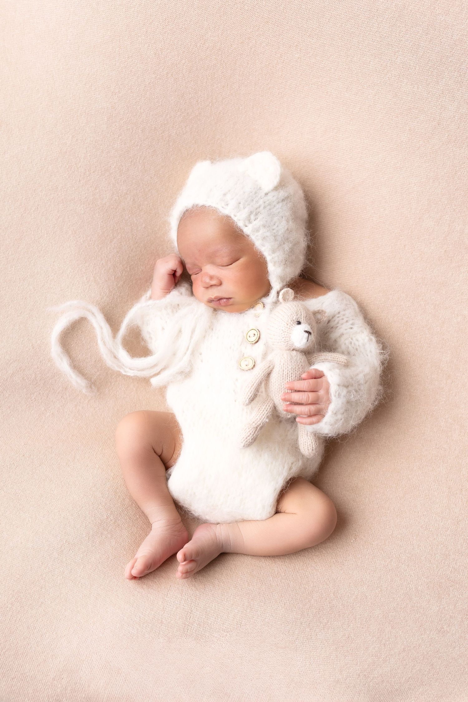 Newborn baby in white knit bear bonnet and romper sleeping while holding a tiny teddy bear. séance naissance chavanoz lyon